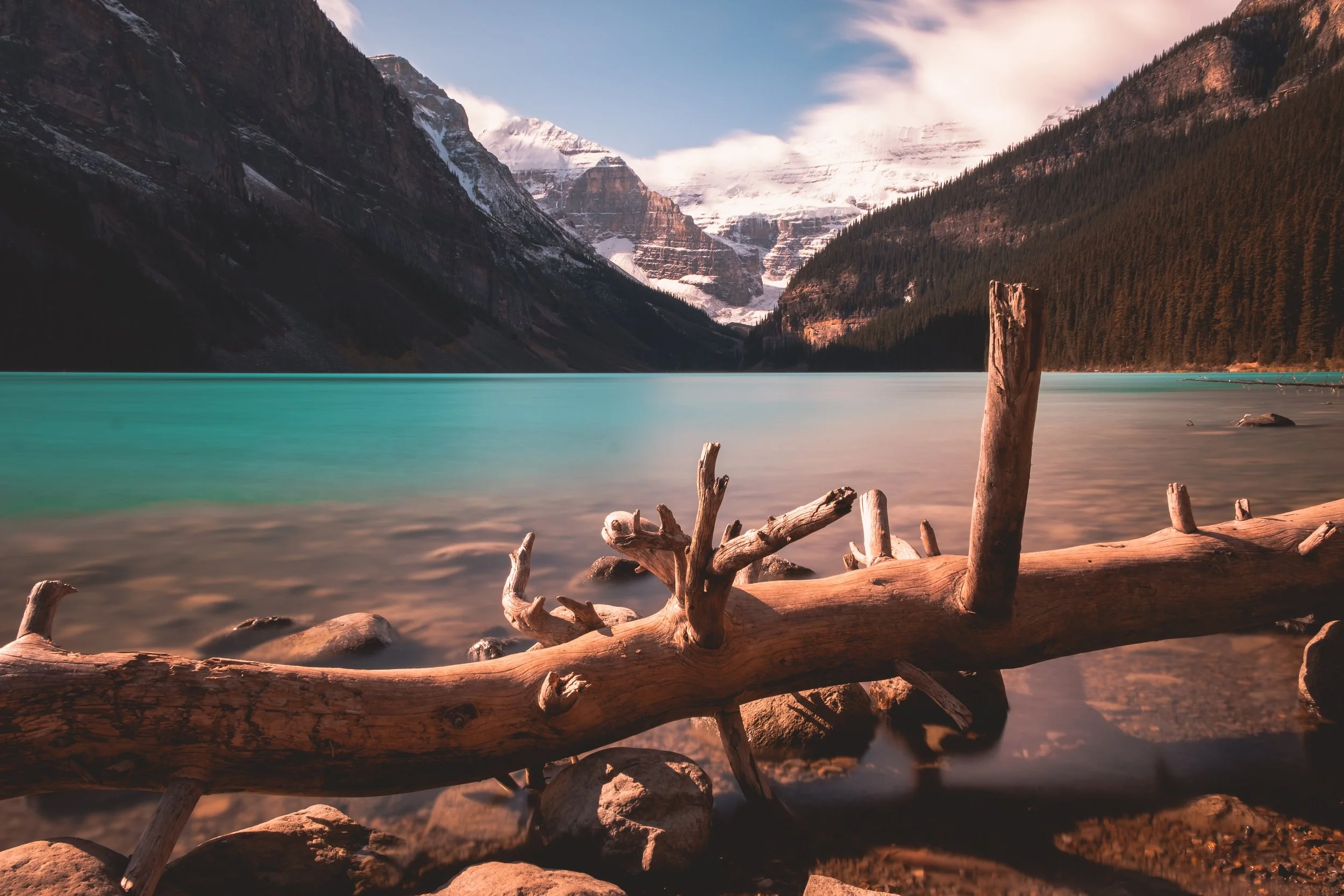 A tranquil mountain lake with turquoise water, surrounded by tall mountains with snow caps, and a fallen log in the foreground on a rocky shoreline.