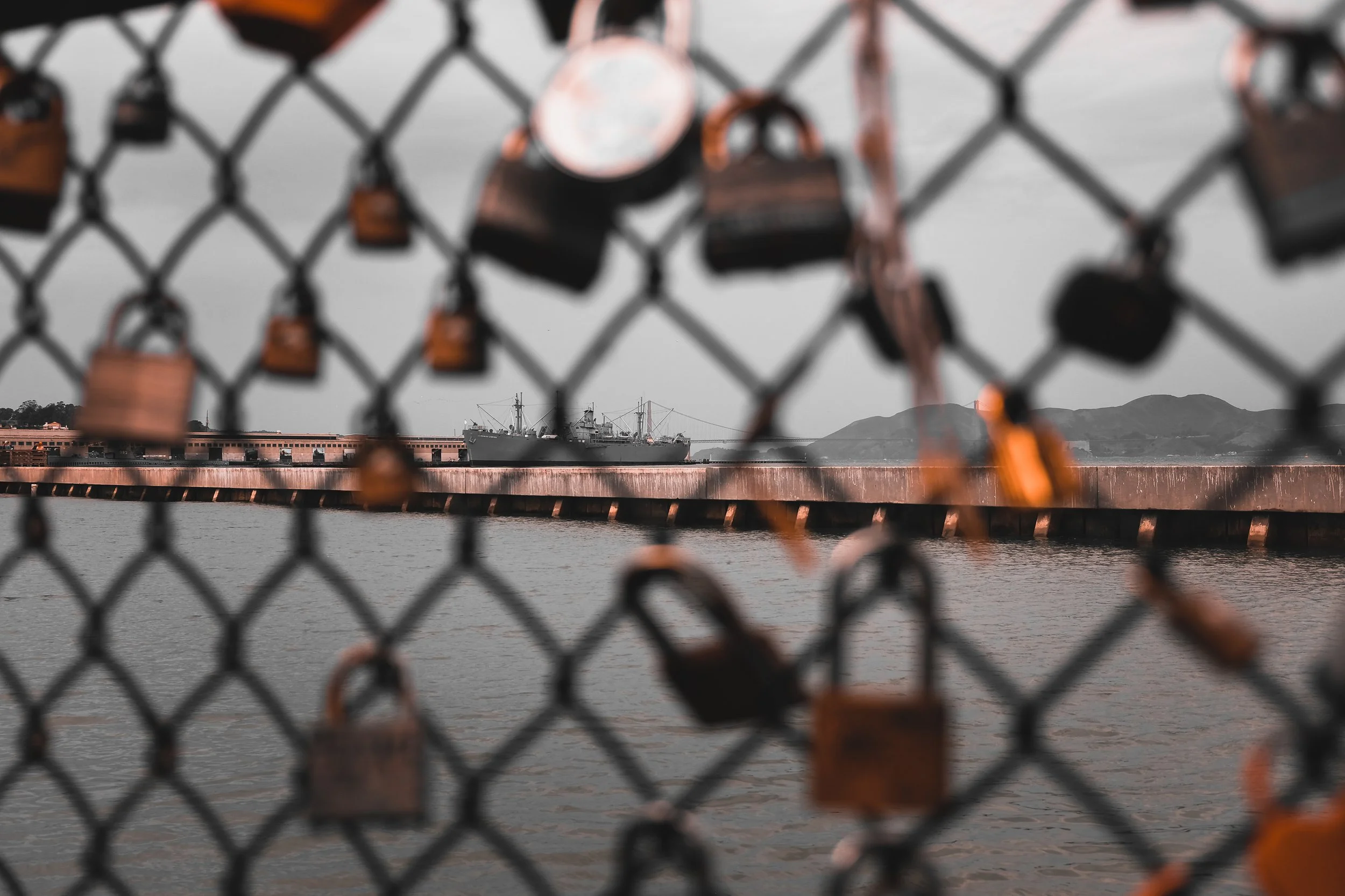 A chain-link fence with numerous padlocks attached, overlooking a body of water with a ship and mountains in the background.