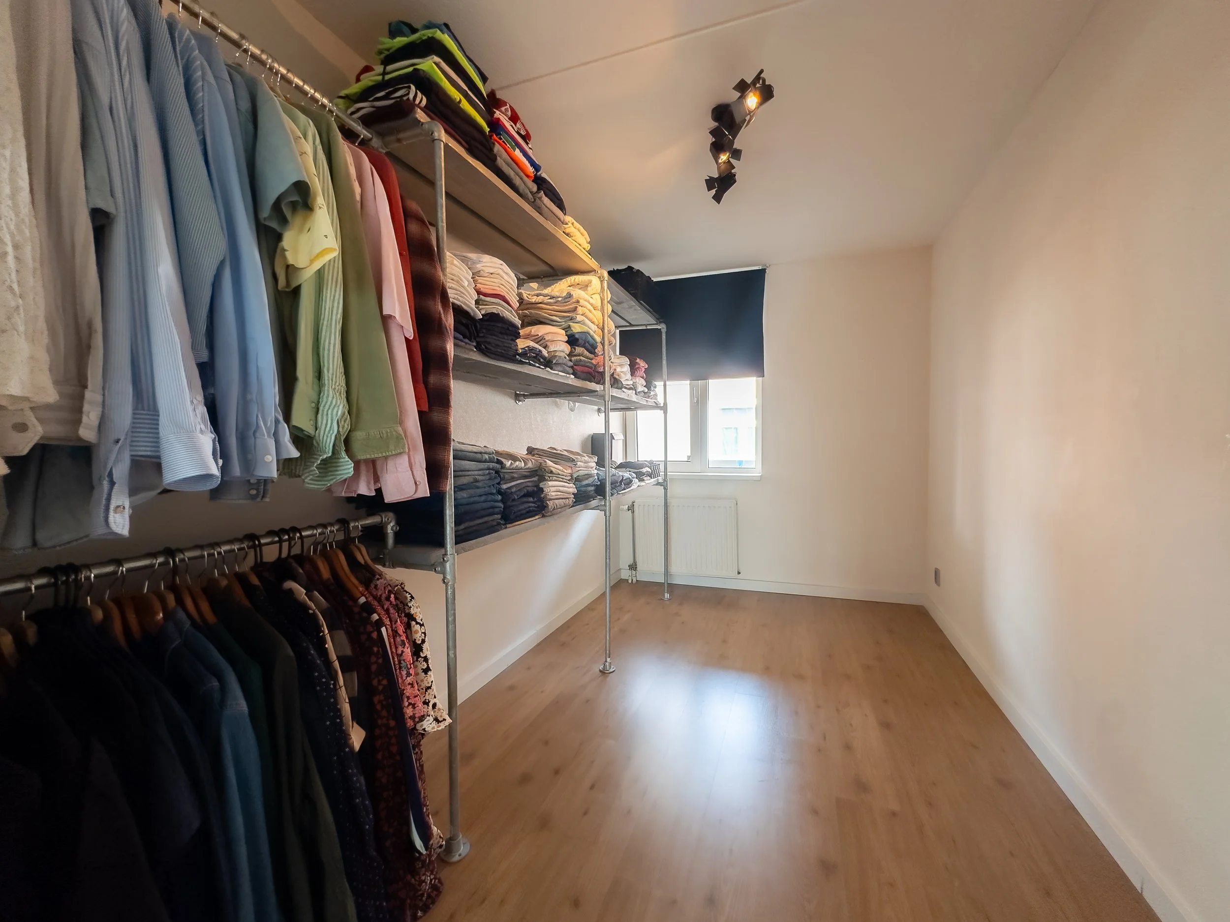 Empty room with clothing racks and shelves filled with folded clothes, a window with black blinds, and wood flooring.