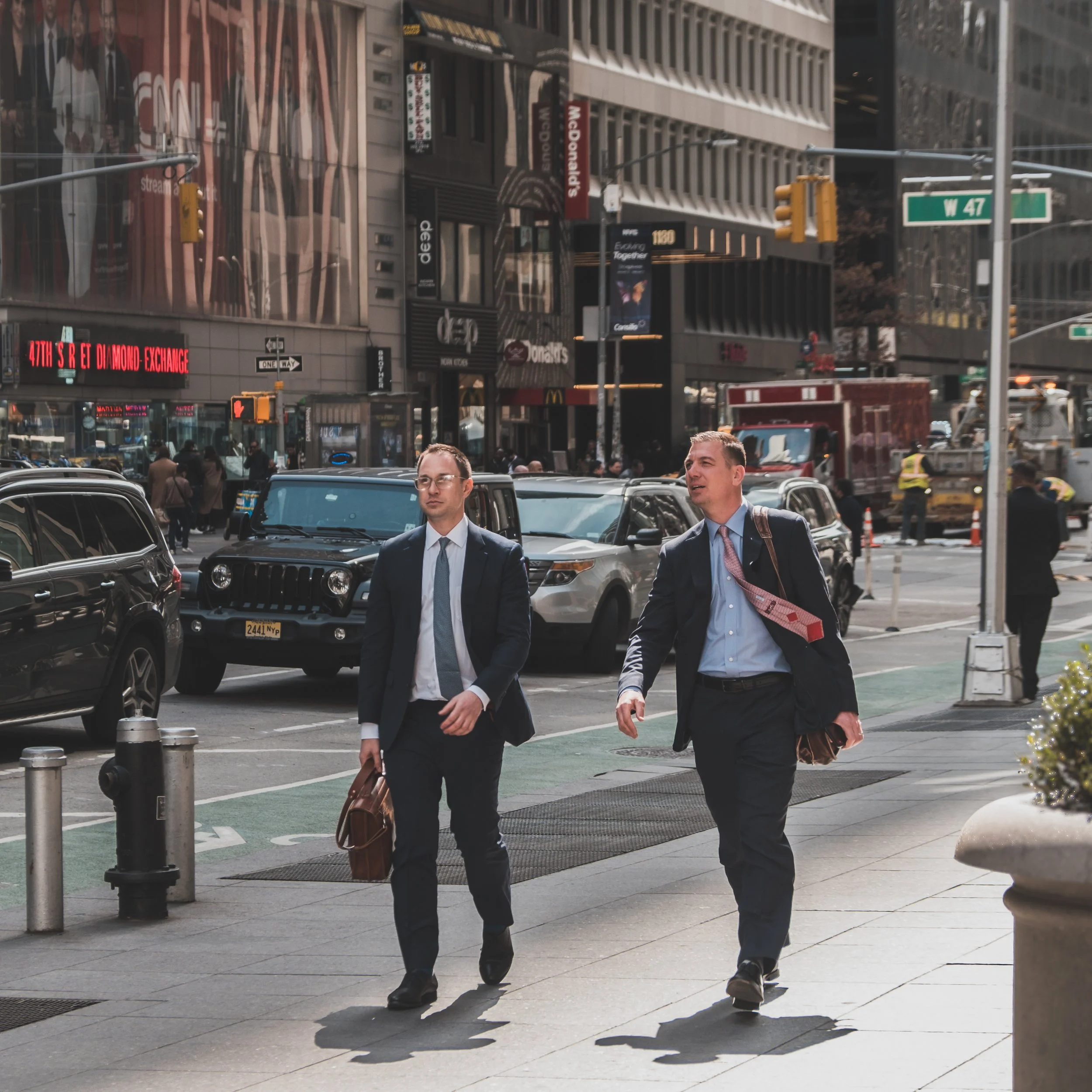 Two men in business suits walking on a city sidewalk, carrying briefcases, with cars and buildings in the background.