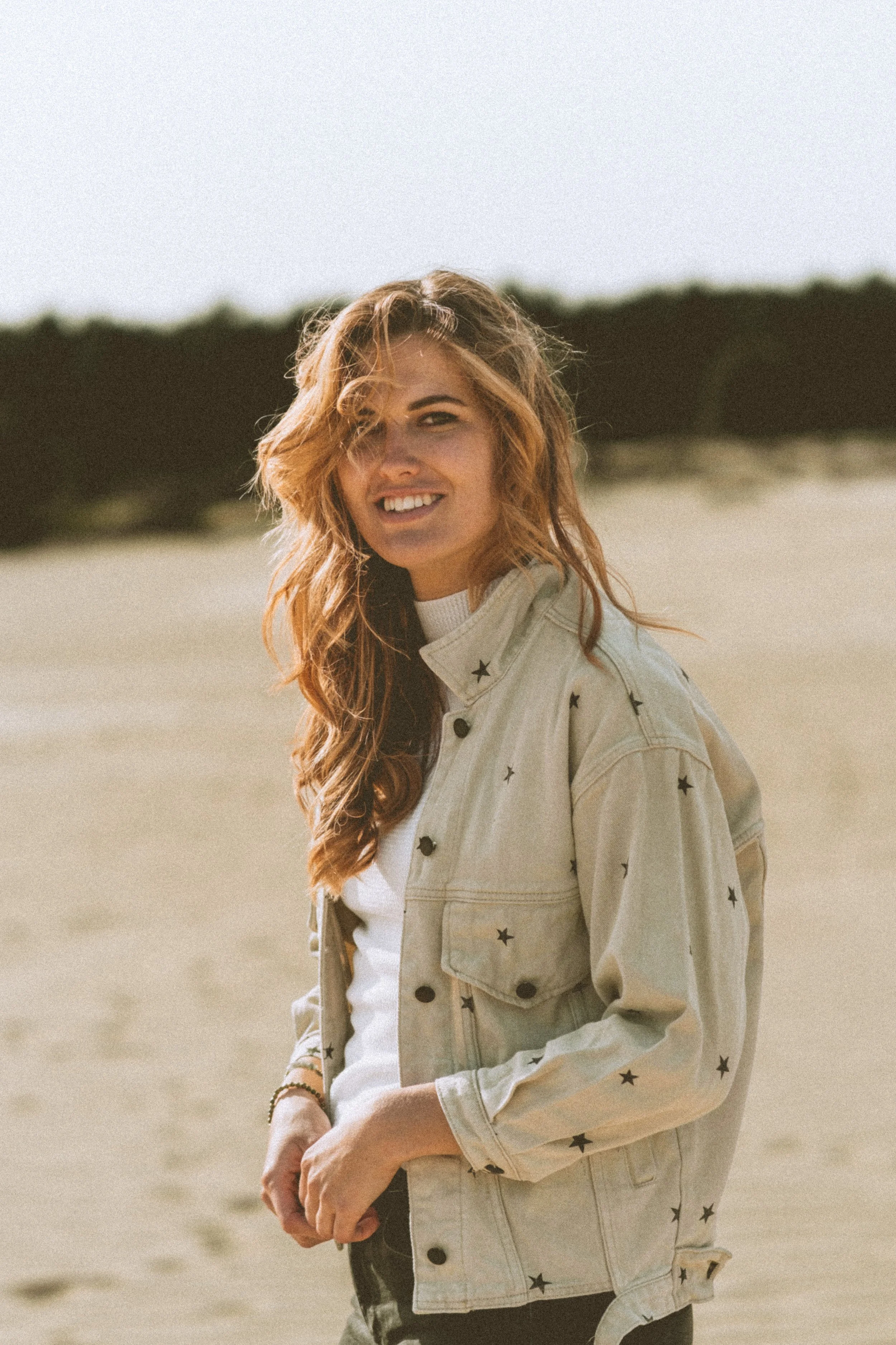 A young woman with wavy hair smiling at the camera, standing on a sandy beach with a blurred background of dunes and sky, wearing a light-colored star-printed jacket.