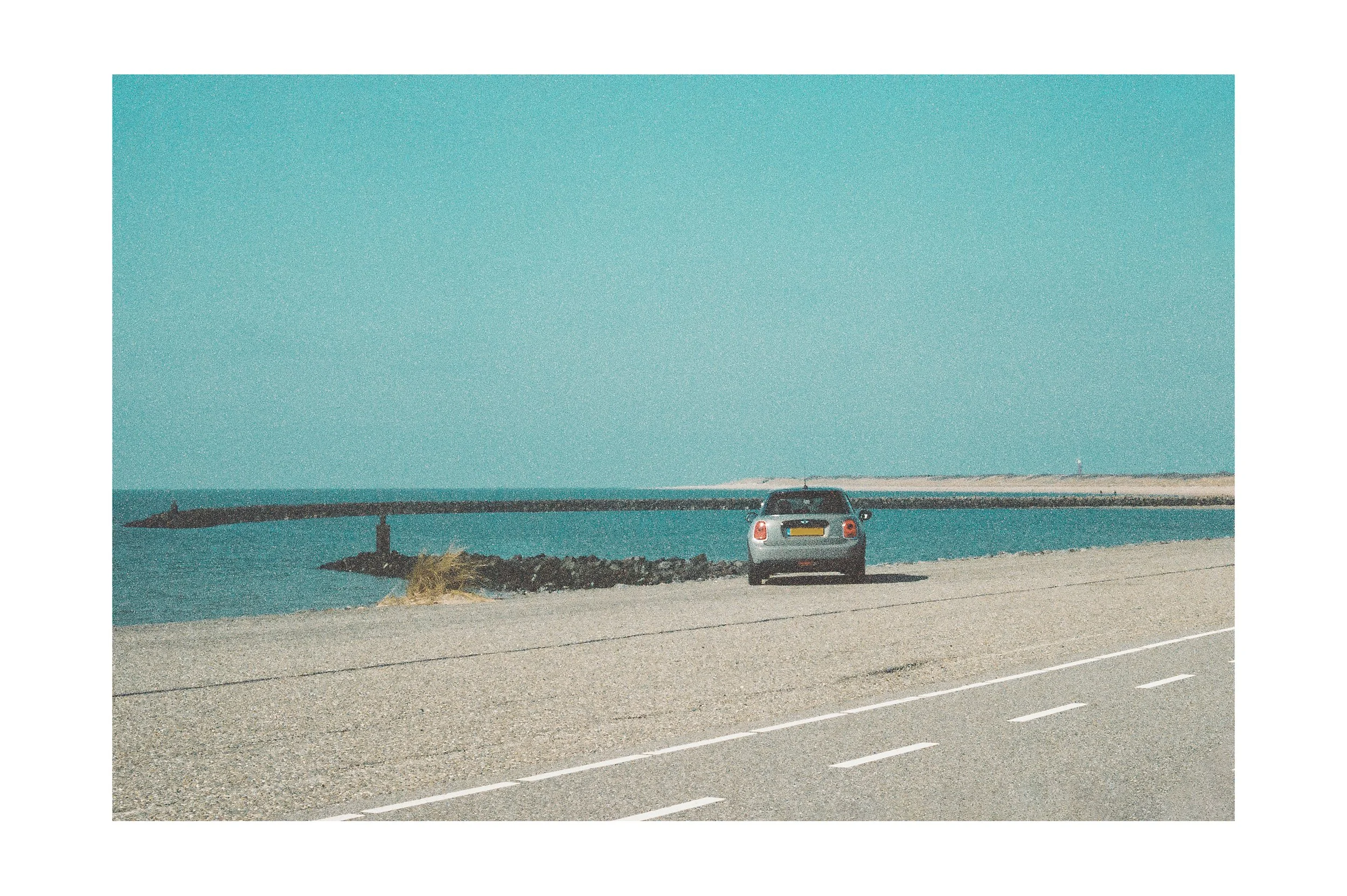 A car parked on a coastal road near the ocean with a breakwater and clear blue sky in the background.