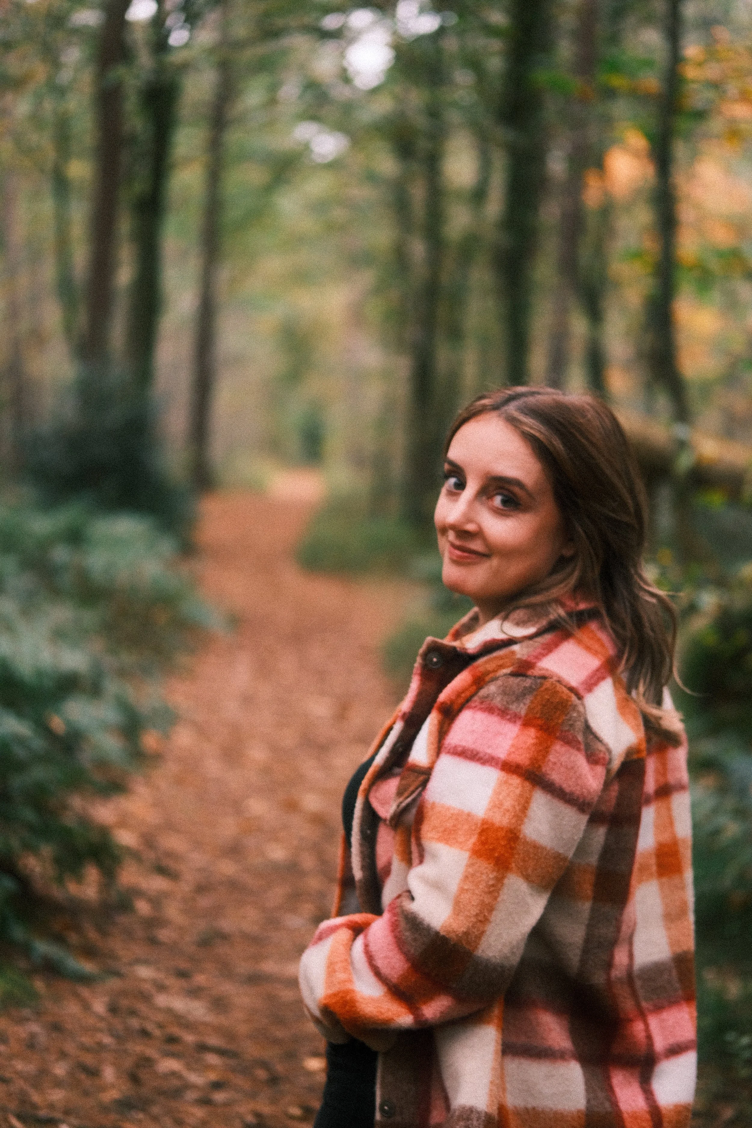 A woman standing on a wooded trail in autumn, wearing a plaid jacket, smiling and looking at the camera.
