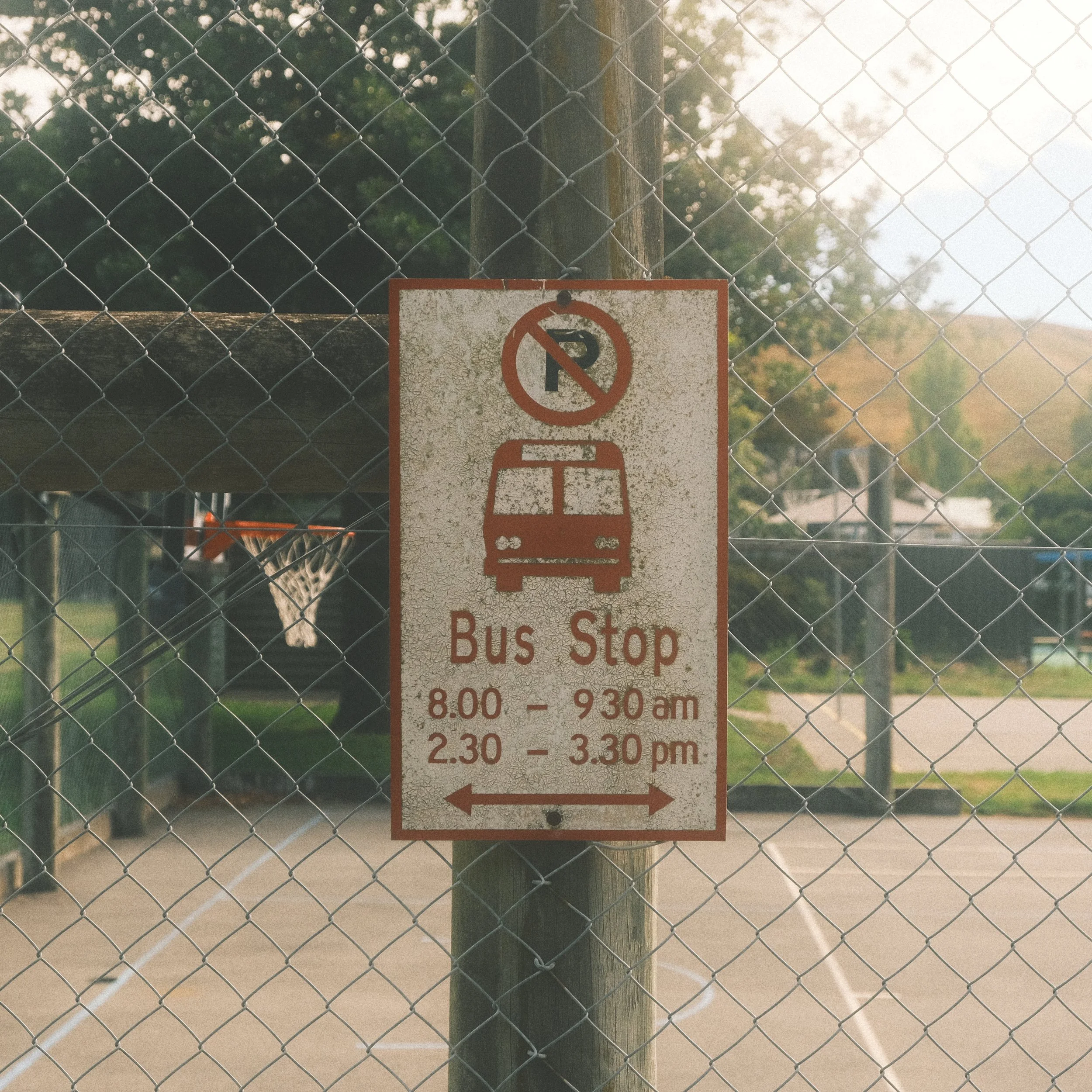 A weathered sign on a chain-link fence indicating the bus stop locations and hours: 8:00 to 9:30 am and 2:30 to 3:30 pm.