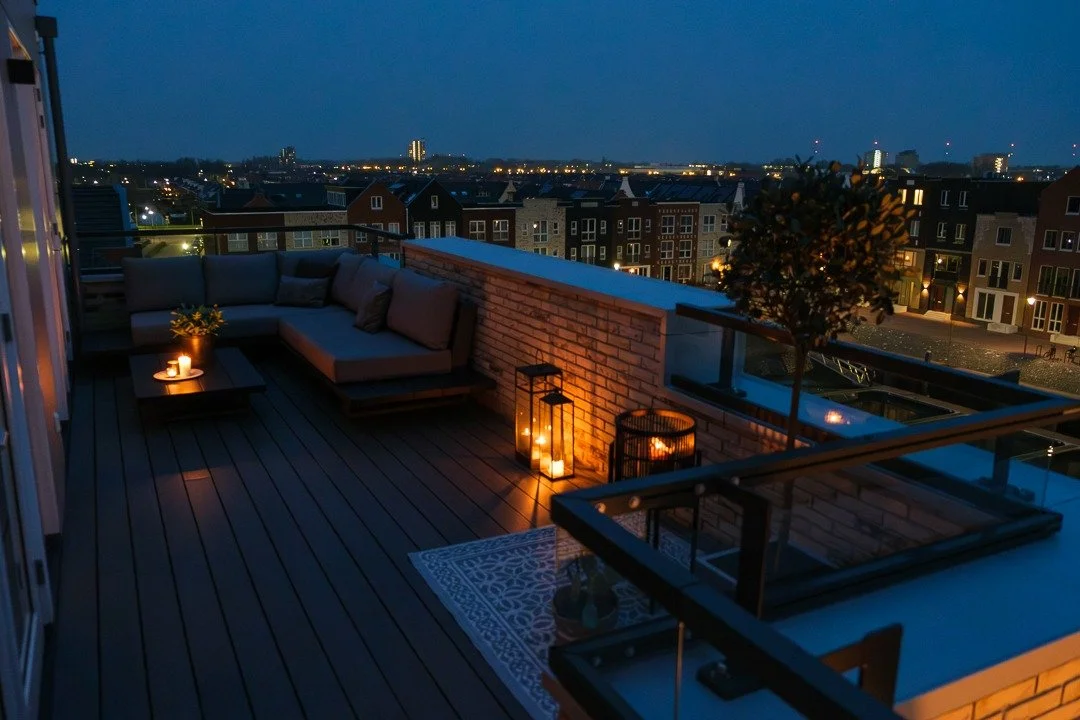 Cozy rooftop balcony at night with outdoor sofa, lit candles, potted plant, and cityscape in the background.