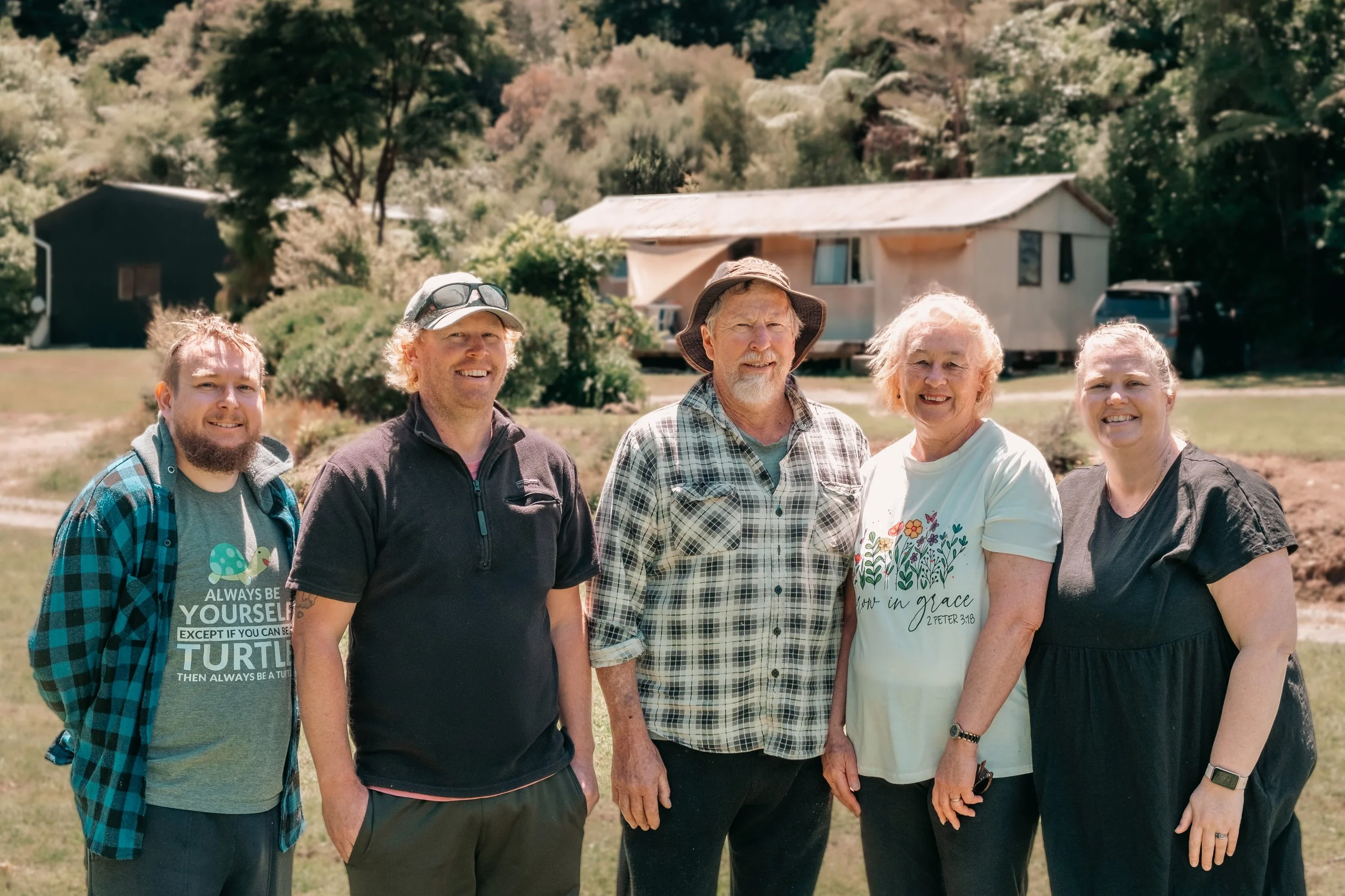 Group of five people standing outdoors on grass, with trees and a house in the background.