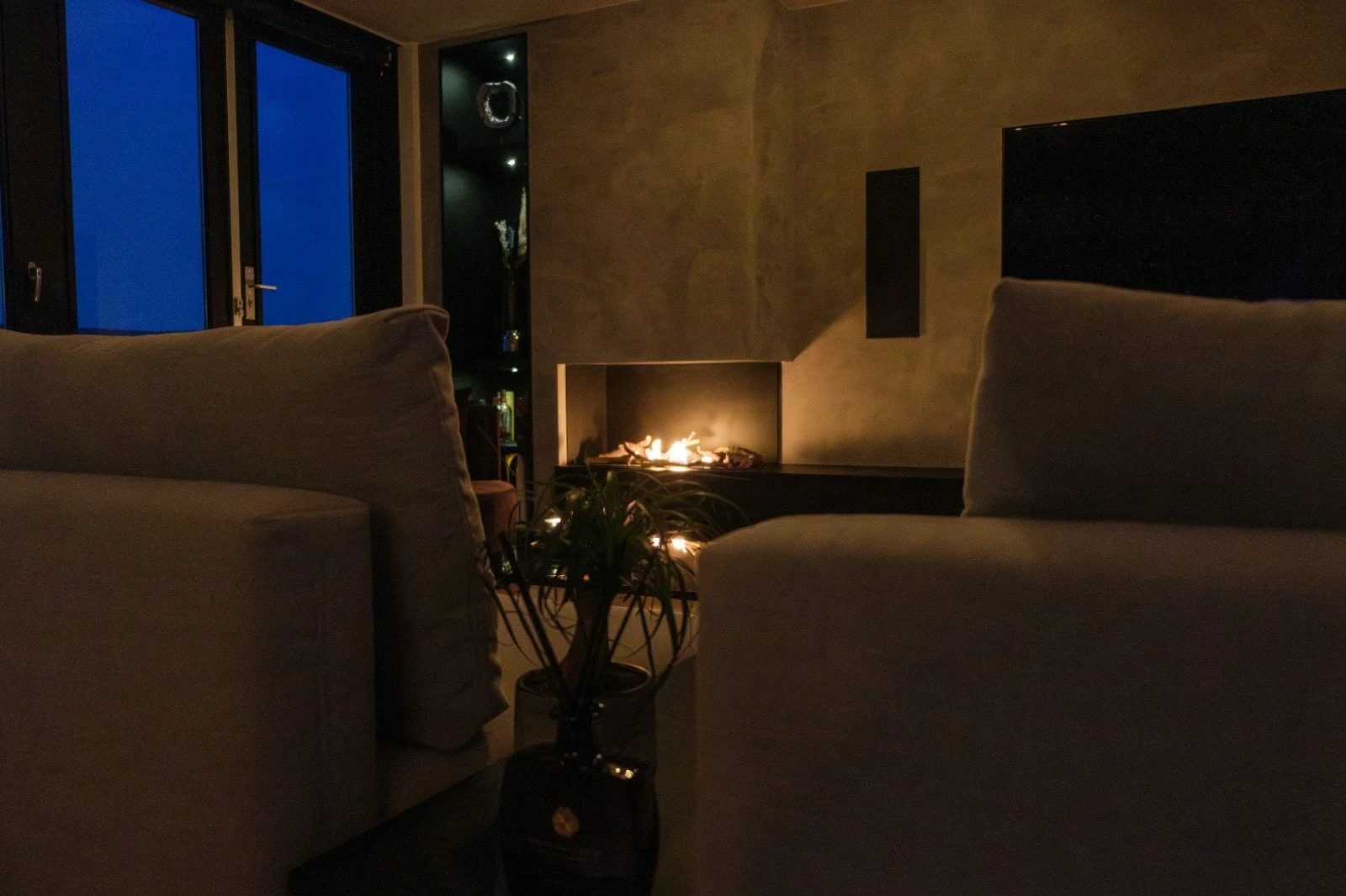 A cozy living room with two beige sofas facing each other, a potted plant on a small table, and a modern fireplace with flames, during the evening or night. There are glass doors leading to a balcony with a view of the blue evening sky.