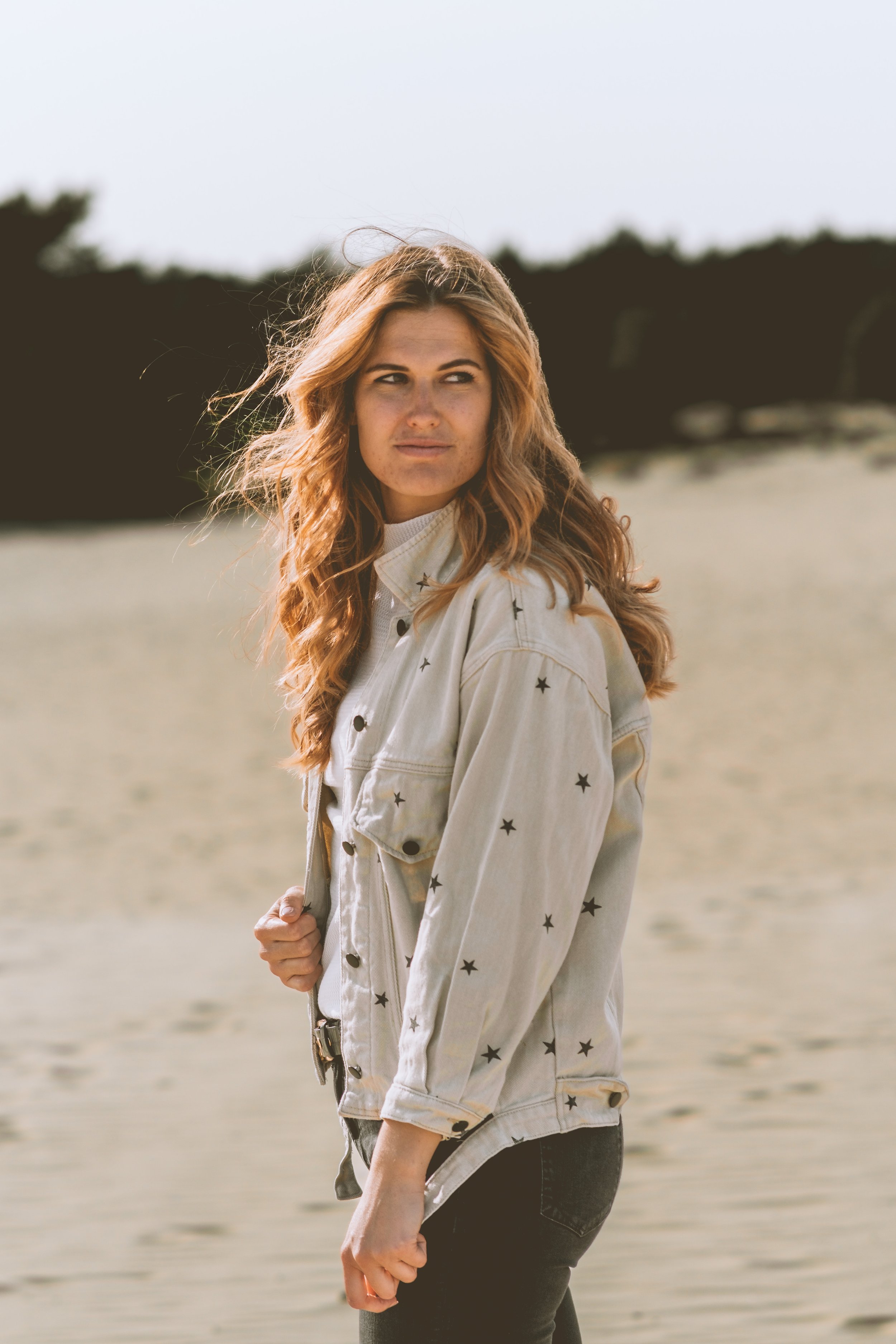 A young woman with wavy, reddish-blonde hair stands near a body of water, looking to her right. She is wearing a light-colored denim jacket with black star patterns and black pants, with a blurred natural background.