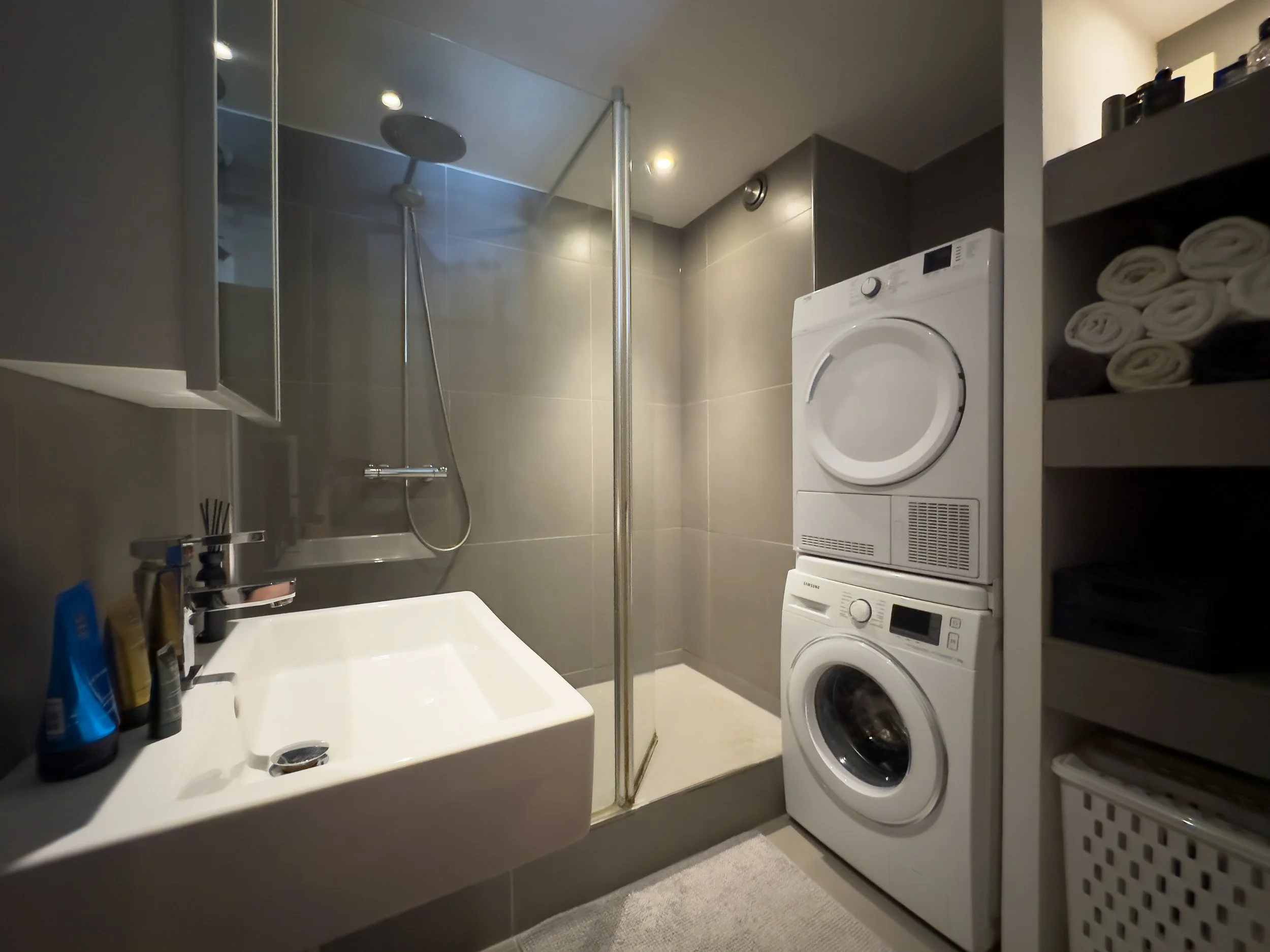 Compact laundry bathroom with a washing machine and dryer stacked beside a shower with a glass door, small sink with toiletries, and shelf with folded towels.