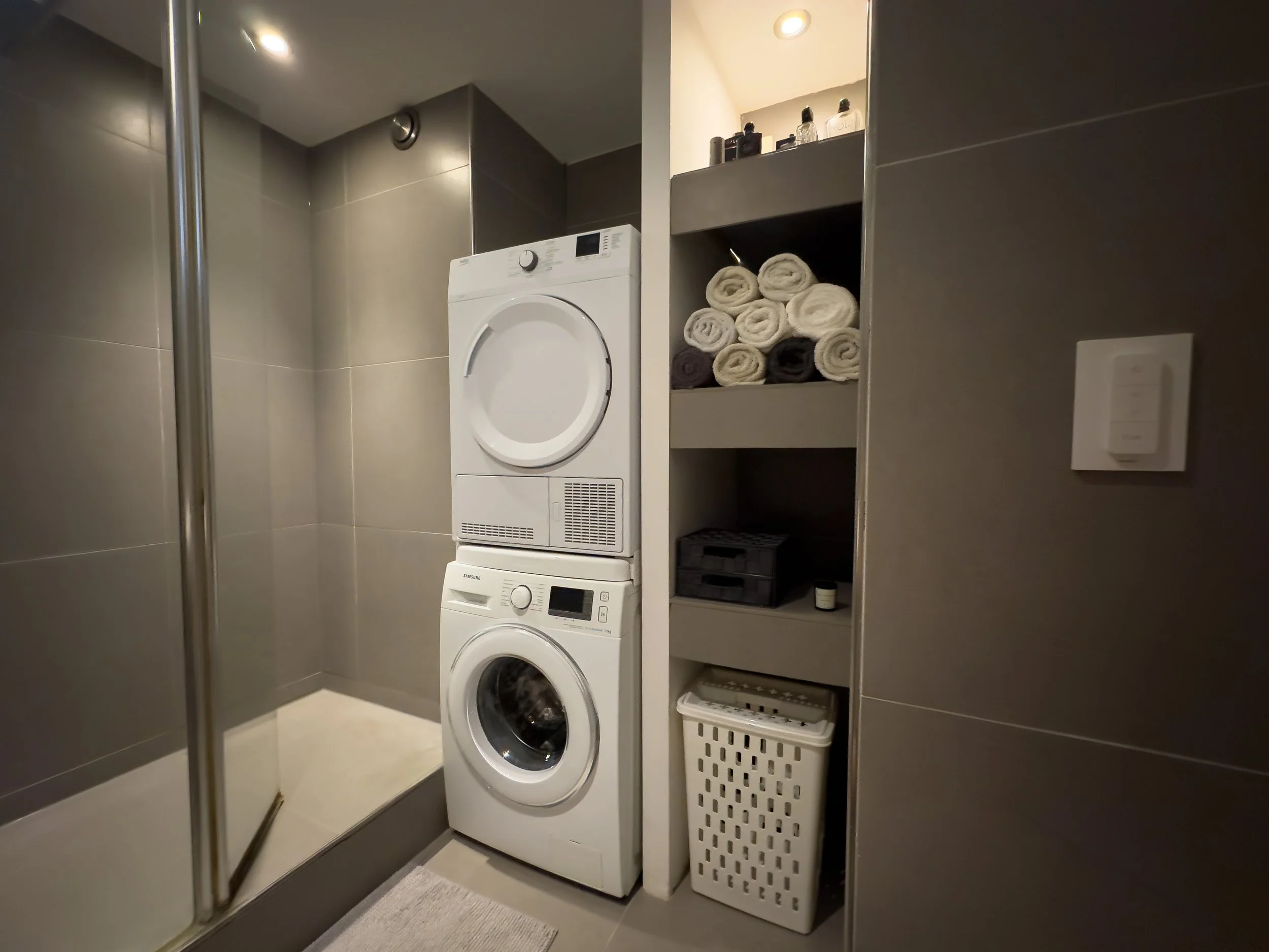 Stacked washer and dryer in a laundry room, with shelves containing rolled towels, small bottles, a black box, a small container, and a laundry basket.