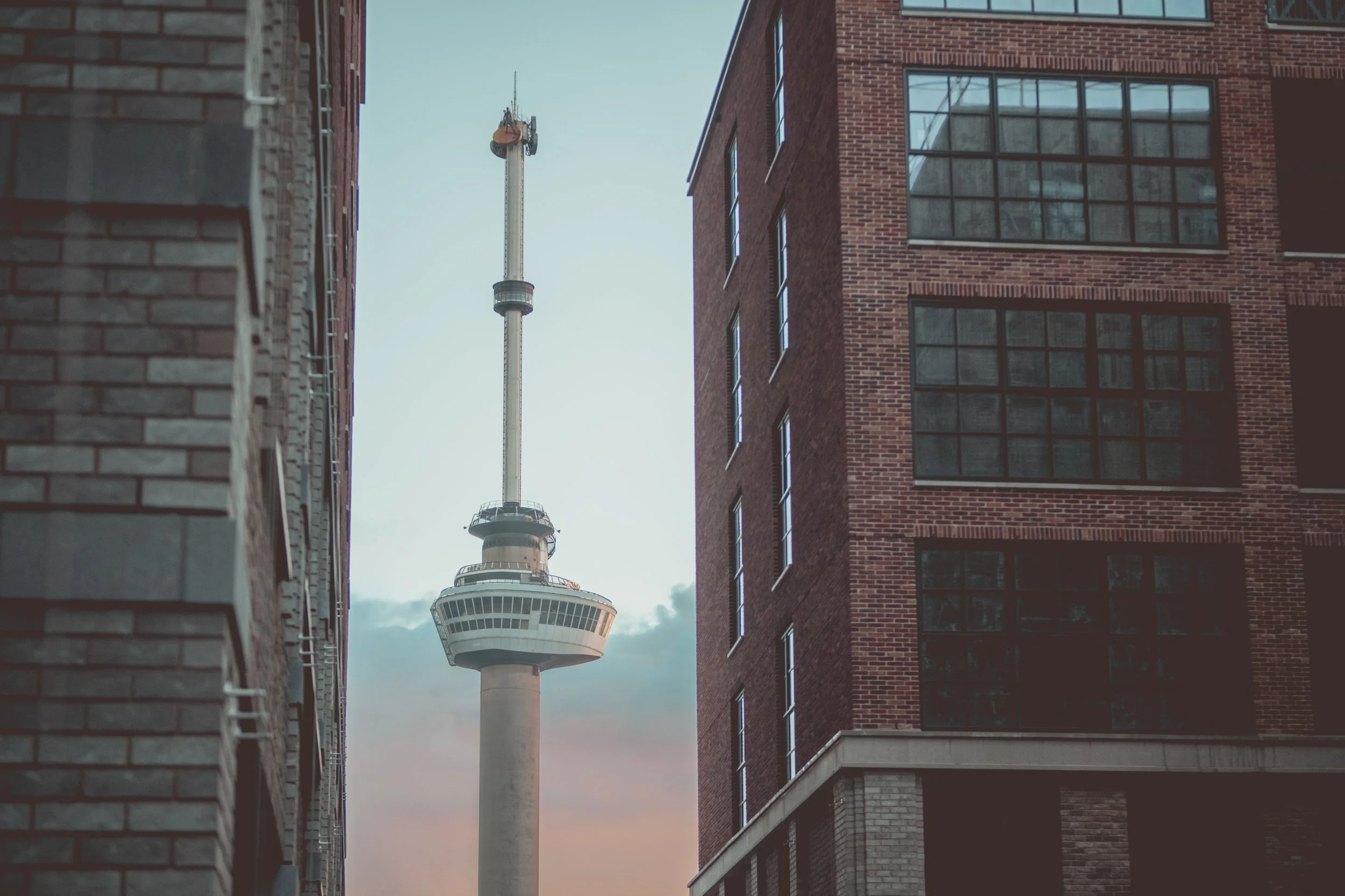 View of a tall communications tower with observation deck, framed by two brick buildings on either side, at dusk in an urban setting.