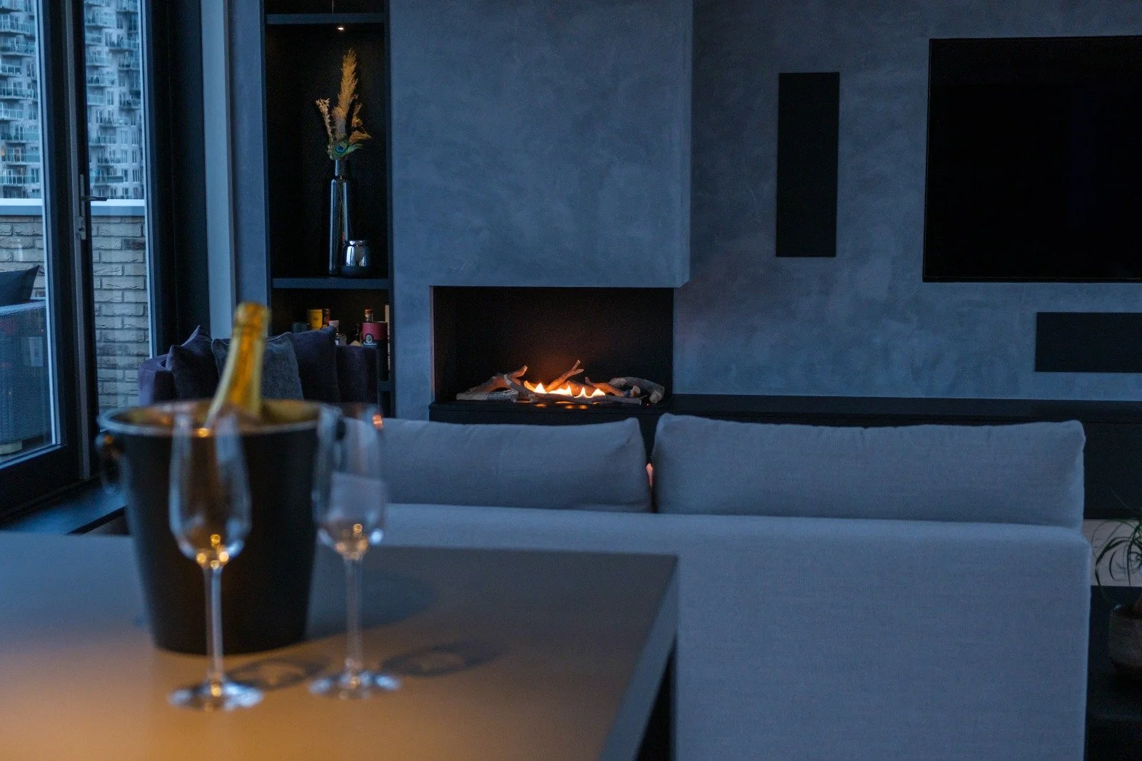Living room with a fireplace, a gray sofa, a dining table with a bucket of champagne and empty glasses, large windows showing a cityscape, and minimal modern decor.
