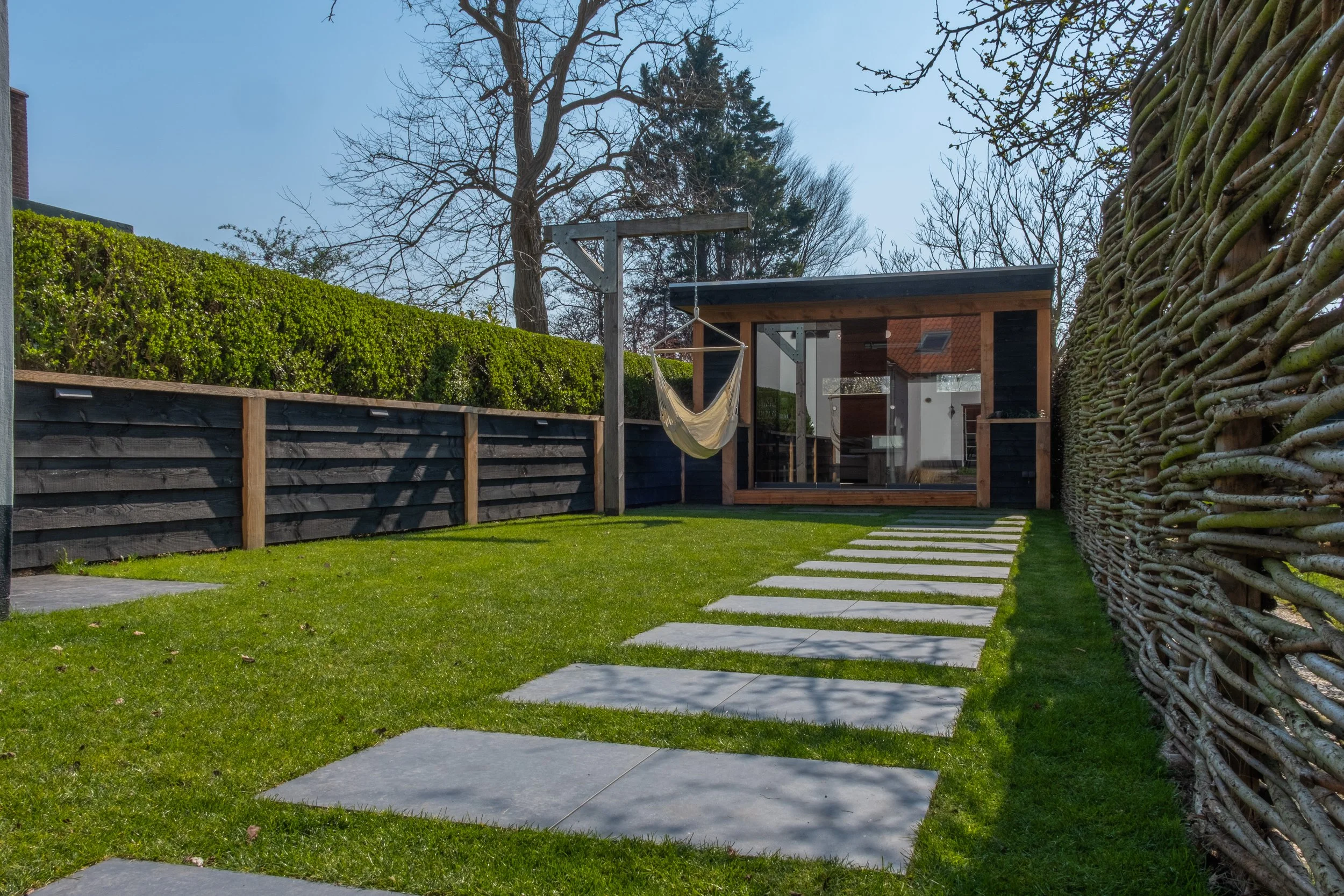 A backyard with a grassy lawn, stepping stones leading to a modern wooden and glass shed, a hammock hanging, a hedge, and a woven fence on the right side.