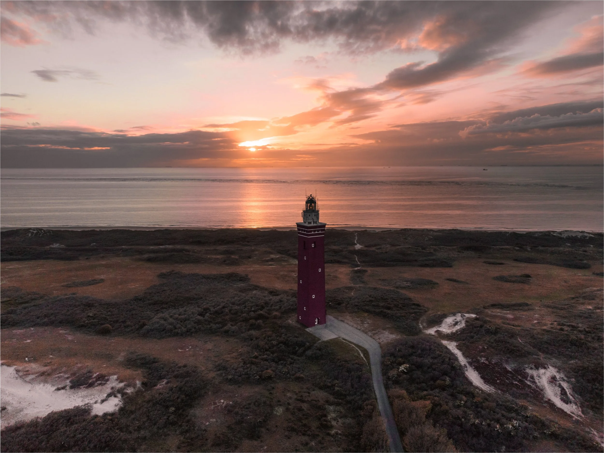 A tall red lighthouse on a coastal landscape at sunset, with a partly cloudy sky and the ocean in the background.