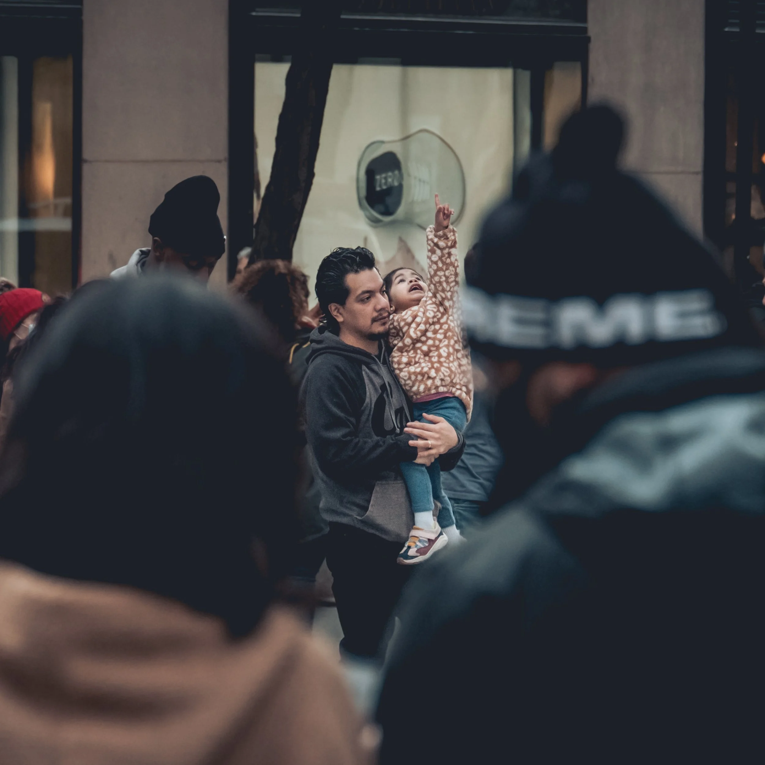 A man holding a young girl in a crowded outdoor setting, the girl pointing upward, surrounded by others.