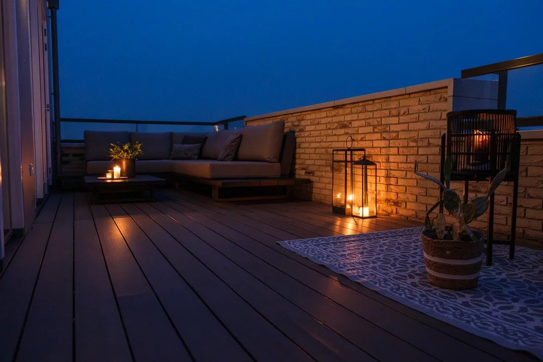 Outdoor rooftop patio at dusk with a gray sectional sofa, candles, lanterns, a patterned rug, and potted plant against a brick wall.