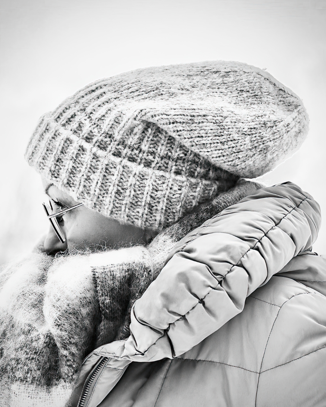 A close-up black-and-white photo of a person wearing a knit beanie, glasses, and a winter jacket, with the person’s face partially visible from the side.