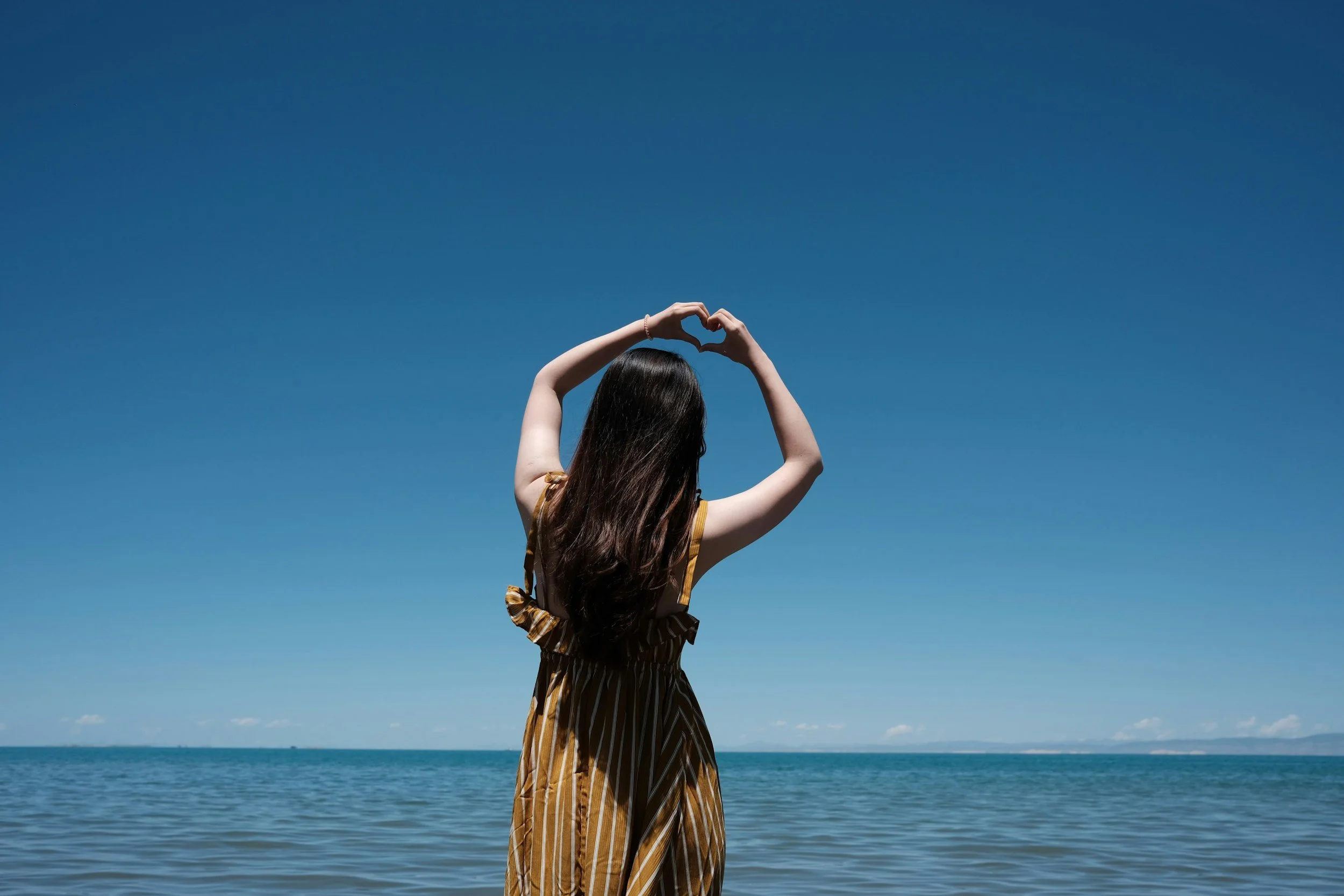 A woman with long dark hair making a heart shape with her hands above her head, standing by the water during daytime with a clear blue sky.