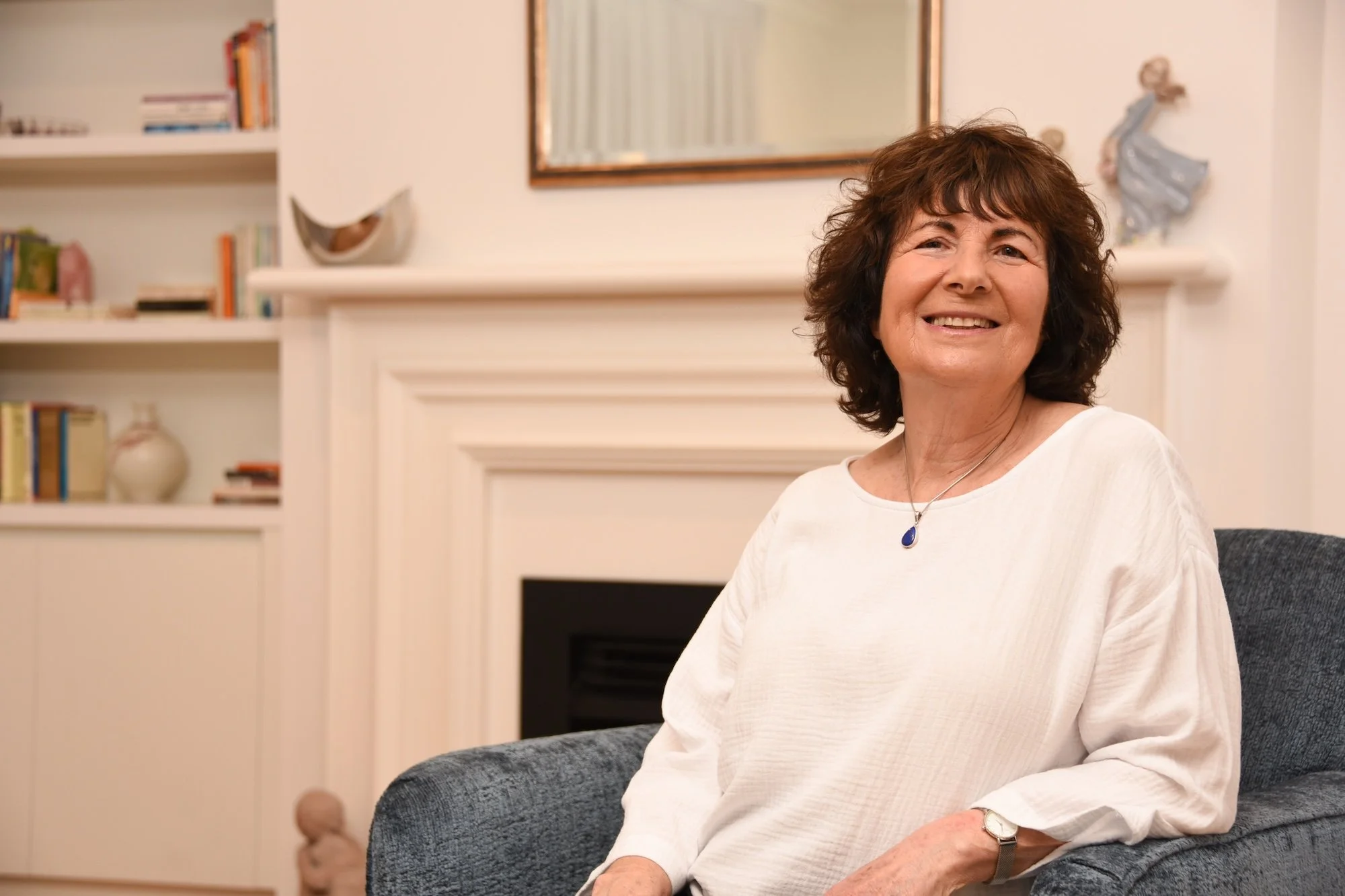 A smiling middle-aged woman with short curly brown hair sitting on a dark gray upholstered chair in a cozy, well-decorated living room. She is wearing a white long-sleeve top and a blue pendant necklace. In the background, there is a white fireplace, a large mirror above it, and white bookshelves filled with books and decorative items on the sides.