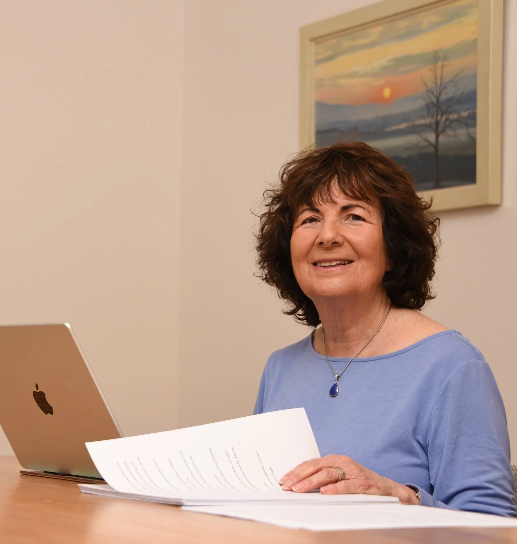 A woman with short brown hair, wearing a blue shirt and a necklace, sitting at a desk and looking at documents while using an Apple MacBook laptop.