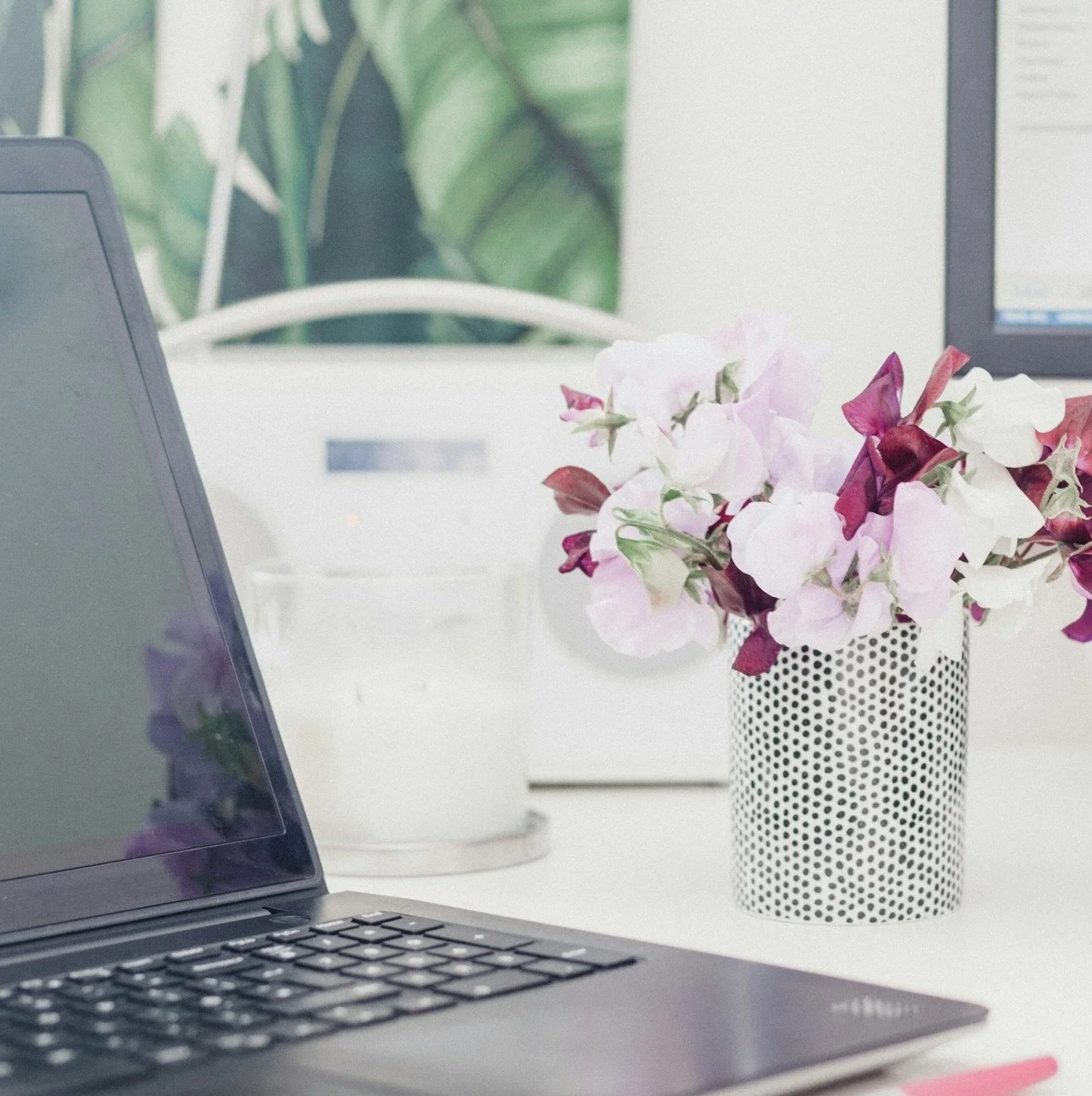A desk with a laptop, a potted plant with pink and white flowers, and a background of a computer monitor and green painting.