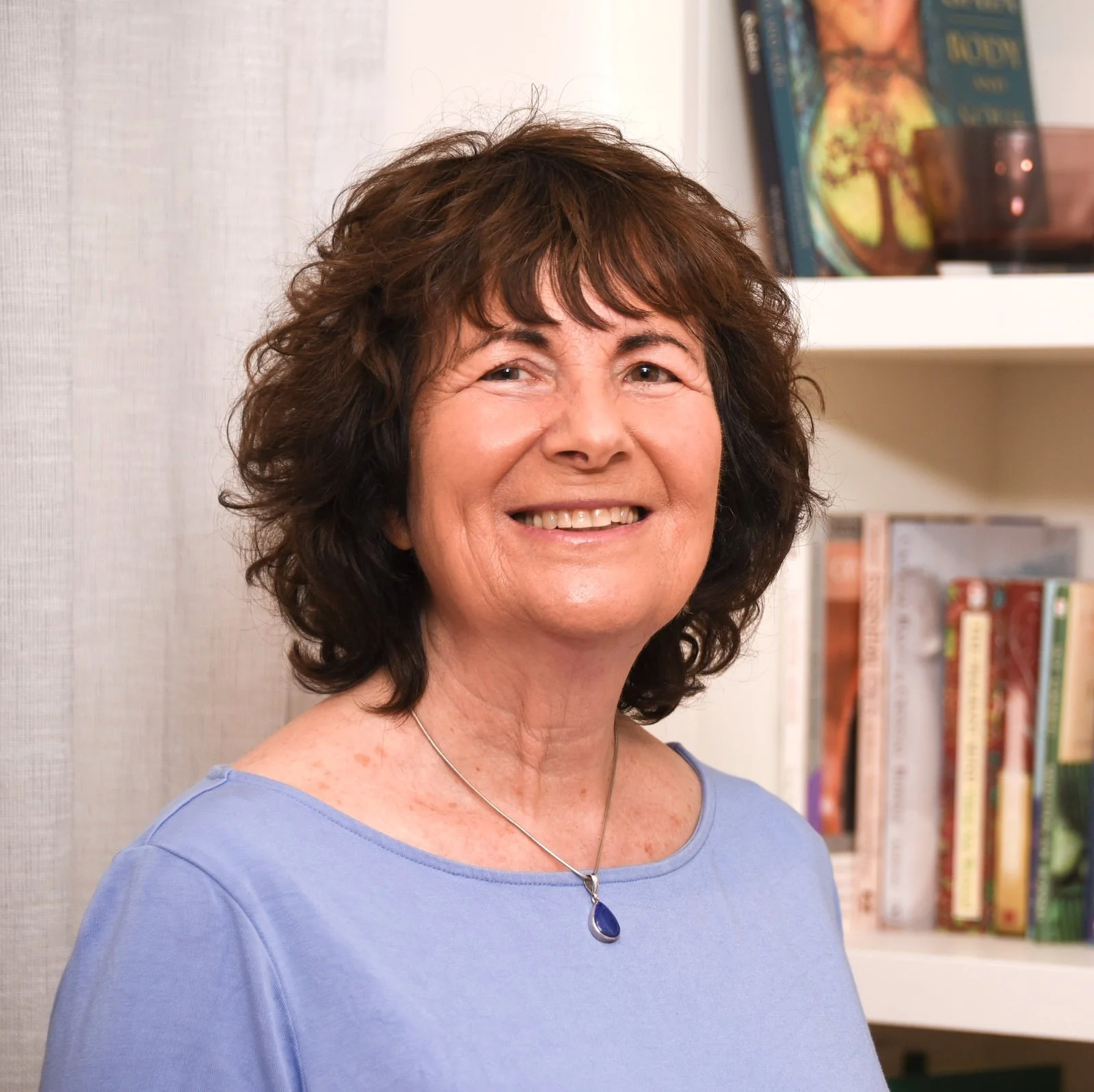 A smiling middle-aged woman with curly brown hair, wearing a light blue top and a silver necklace with a blue gemstone, standing in front of a bookshelf filled with books.