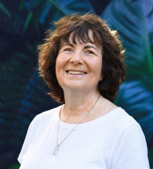 A smiling woman with short, curly brown hair, wearing a white shirt and a silver necklace, standing outdoors with large green tropical leaves in the background.