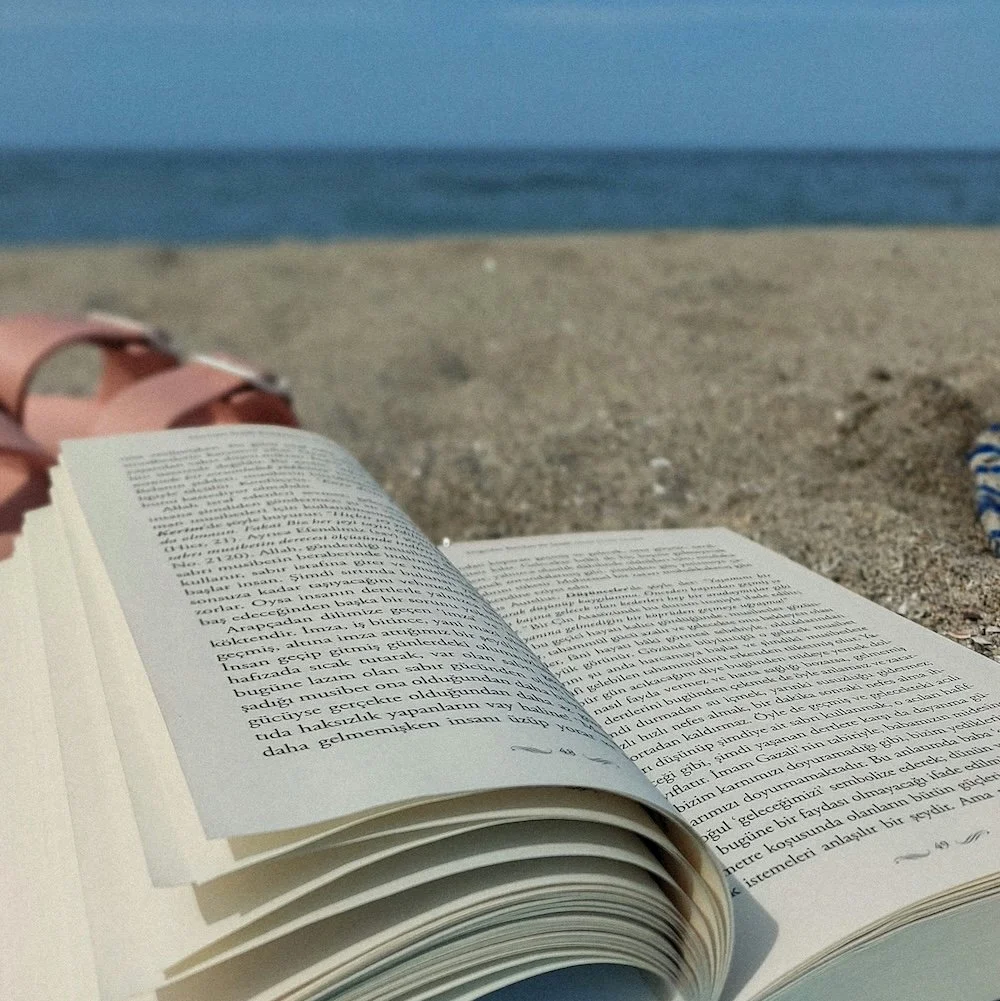 Open book on a sandy beach with ocean waves in the background.