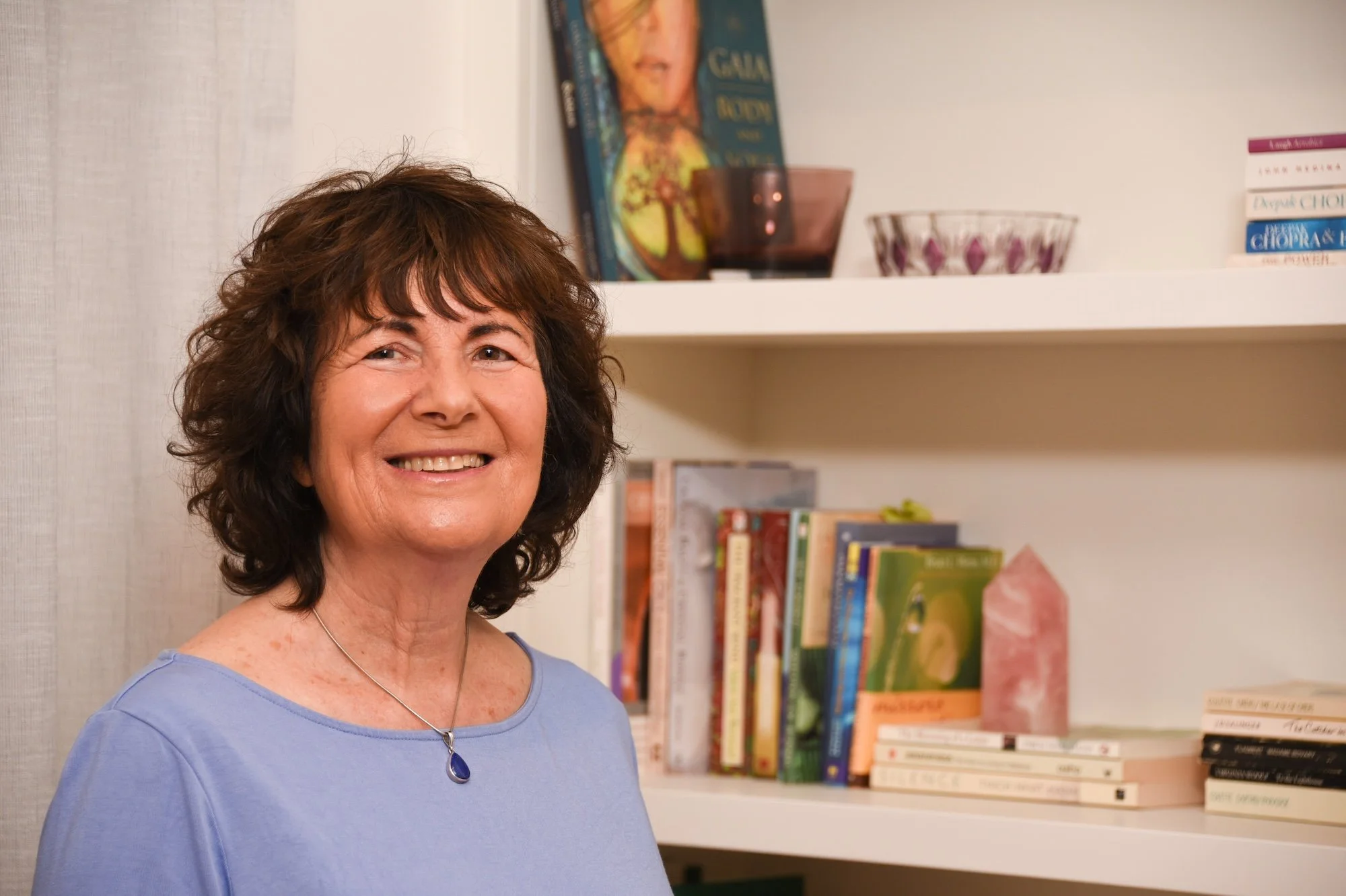 A woman with brown curly hair smiling in front of a bookshelf with books, bowls, and artwork on top.