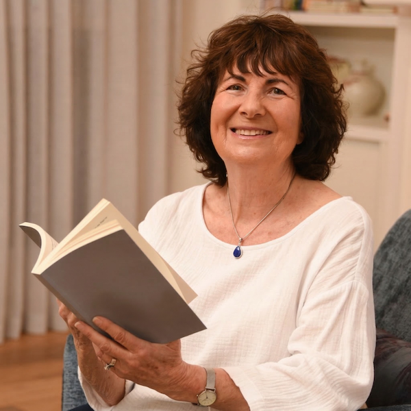 Smiling woman with curly brown hair sitting on a couch, holding an open book, in a cozy room with bookshelves.