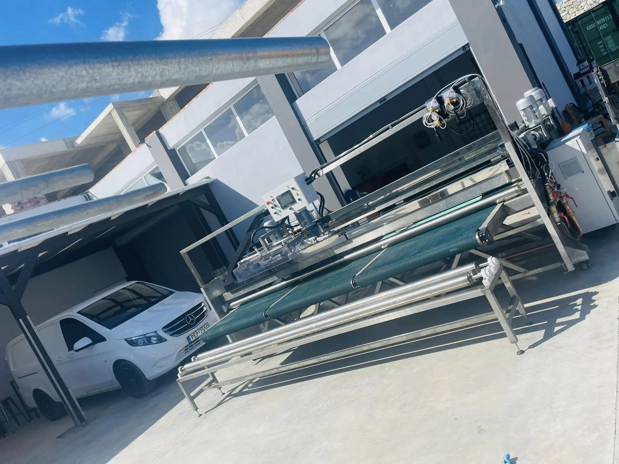Large industrial machine with a conveyor belt, electronic control panel, and various hoses and wires, situated outdoors on a sunny day next to a white Mercedes-Benz van parked in front of a building.