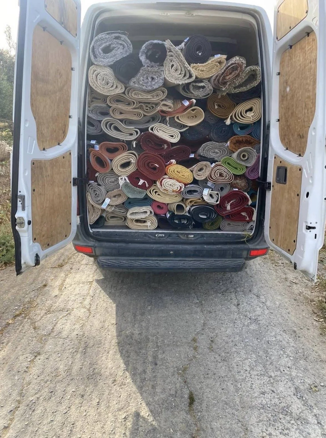 Van with open doors filled with rolled-up carpets and rugs in various colors and sizes, outside on a dirt surface.