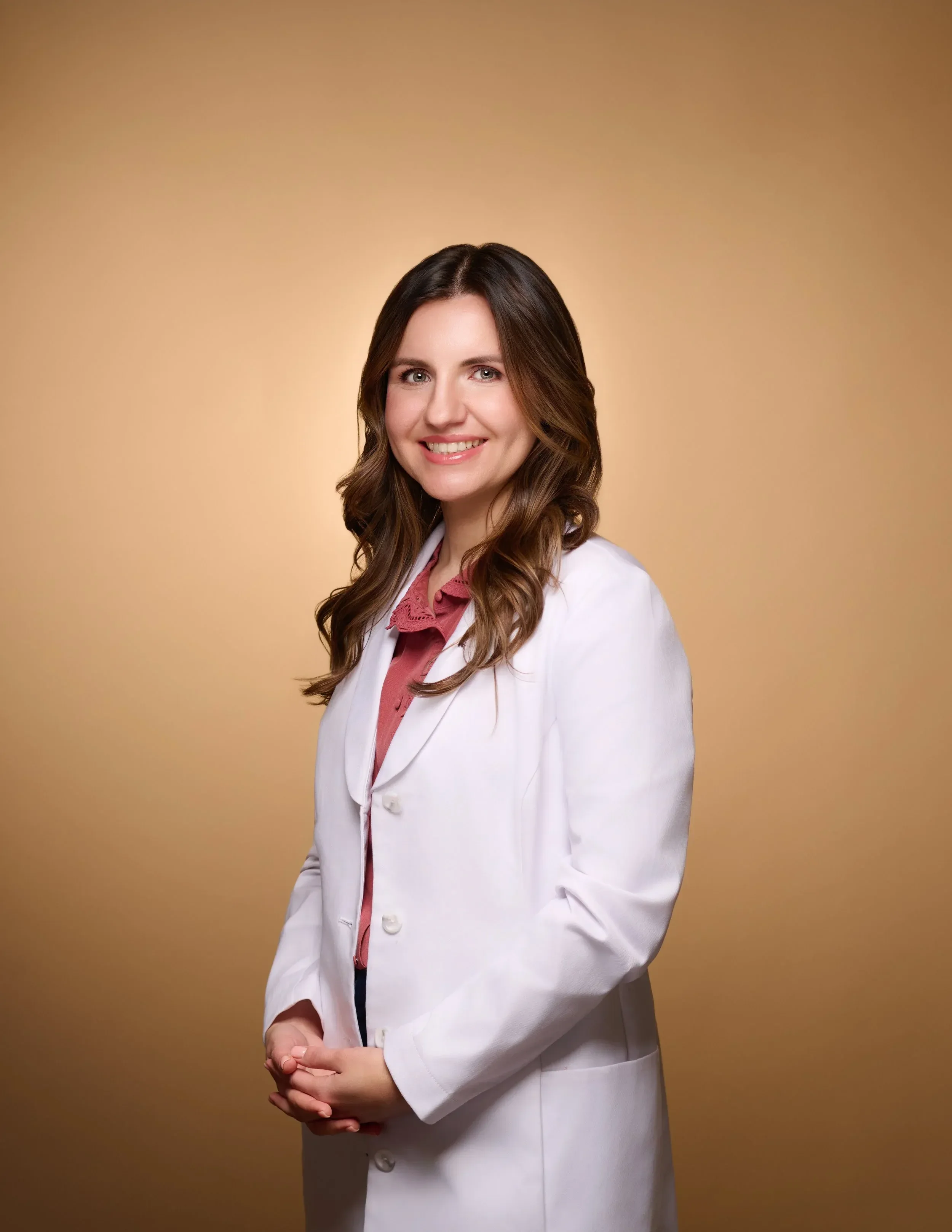 Dr. Myrna Vityuk wearing a white lab coat and a pink shirt, standing against a plain beige background.