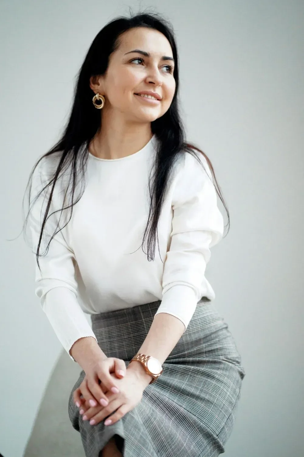 A woman with long dark hair wearing a white blouse and plaid skirt sits smiling, gazing to the side.