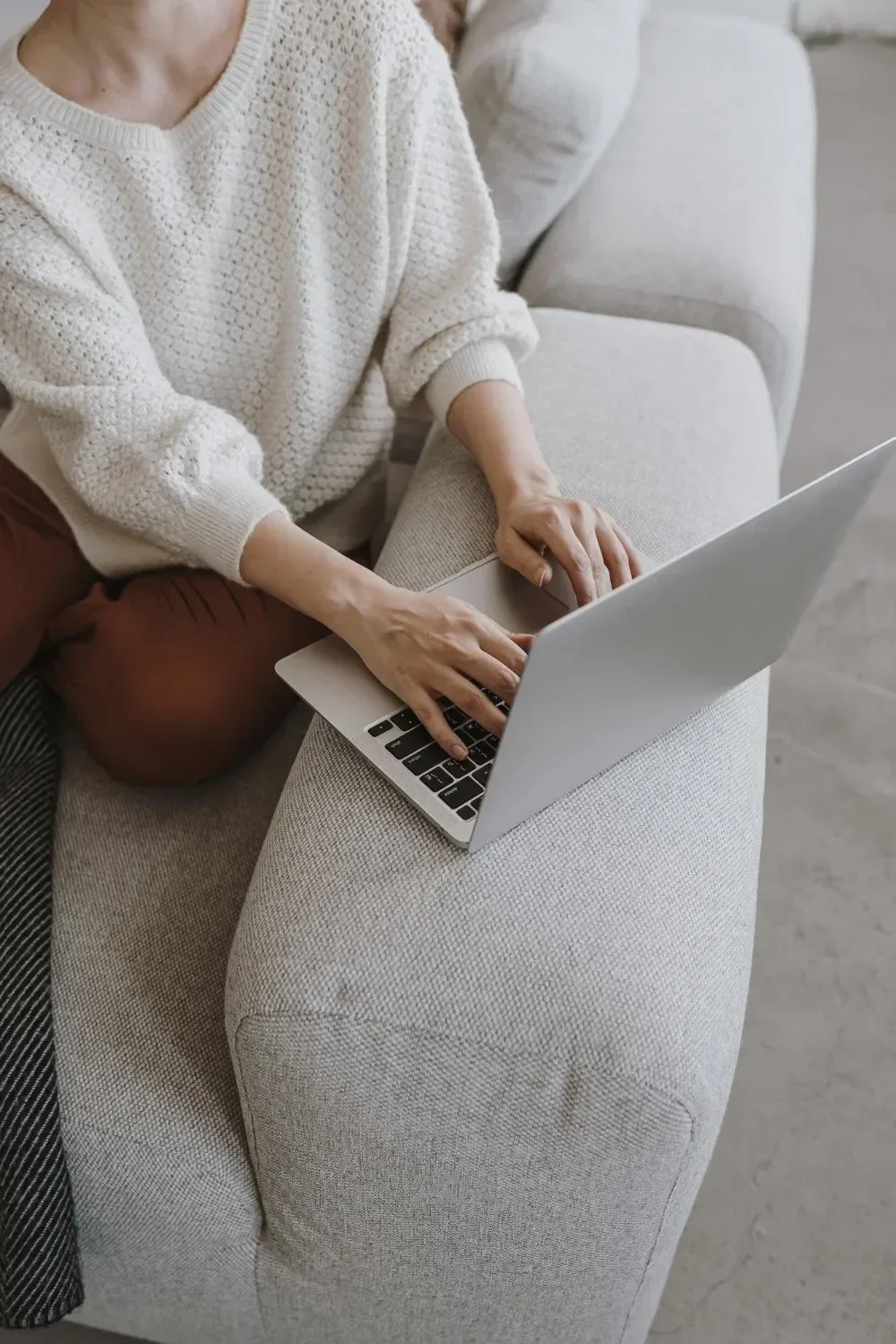 A person in a white sweater types on a laptop while sitting on a light grey sofa.