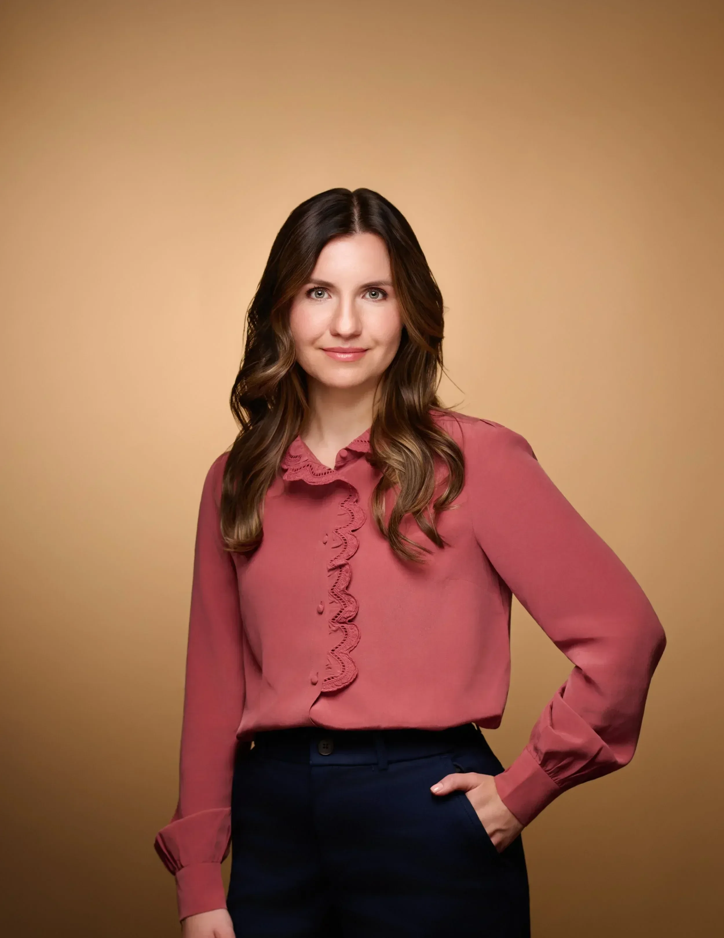 Dr. Myrna Vityuk wearing a pink blouse with lace detailing and dark pants, posing against a beige background.