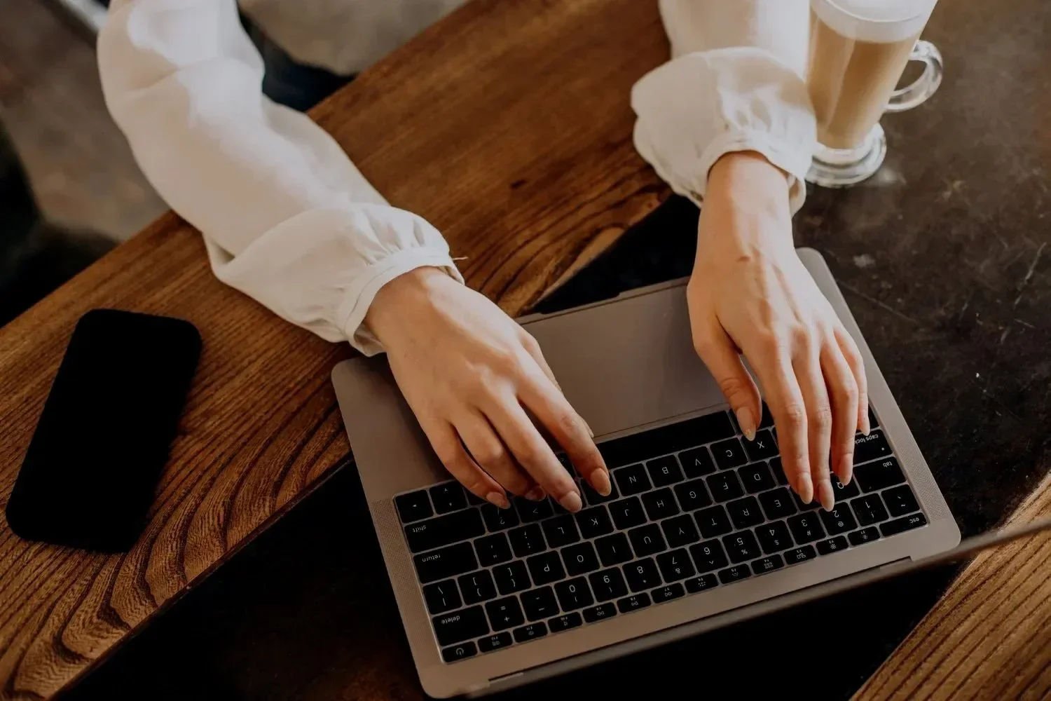 Hands typing on a laptop at a rustic wooden table with a smartphone and a latte nearby, creating a cozy, focused work atmosphere.
