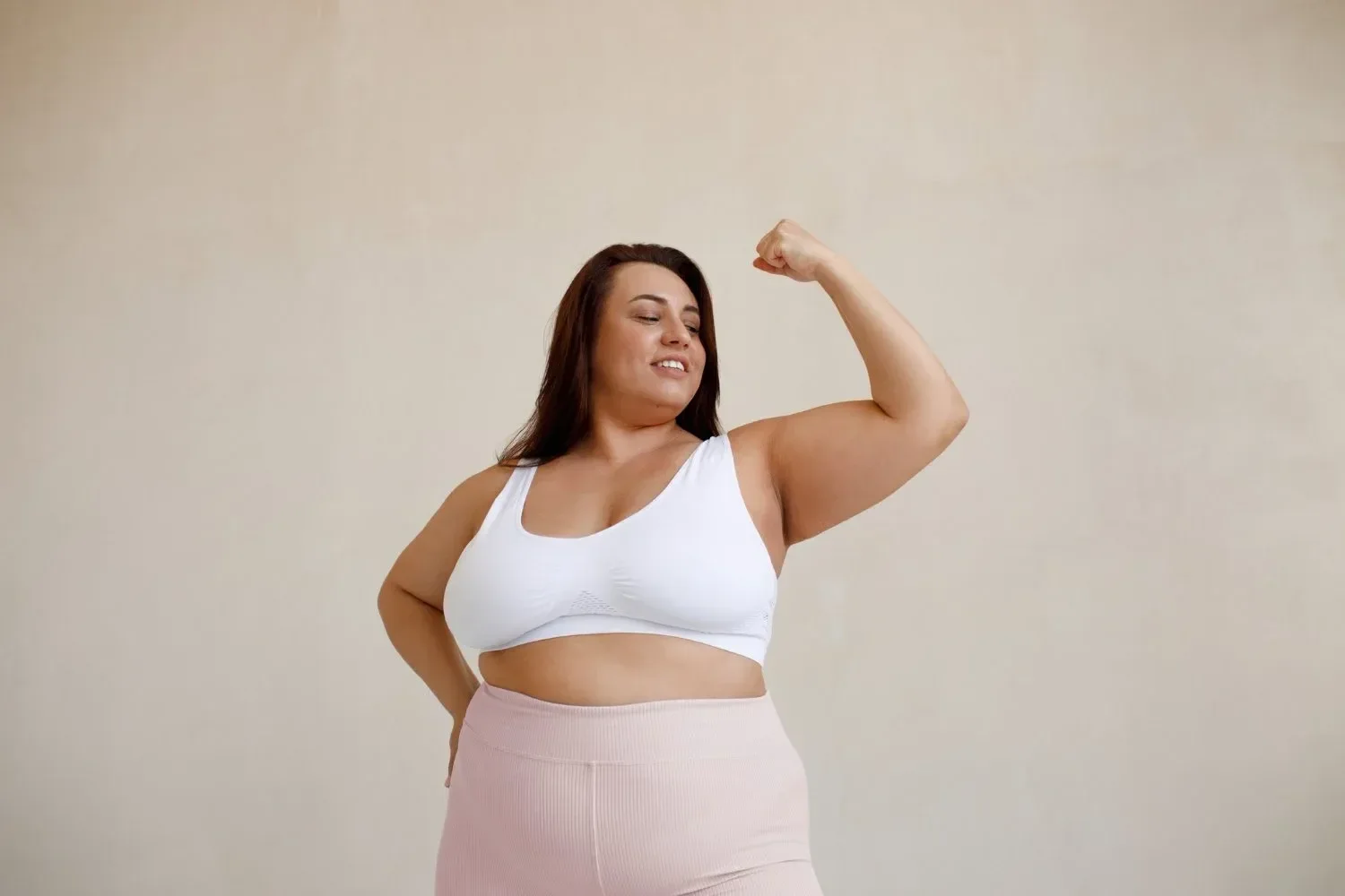 A confident woman in a white sports bra and pink leggings smiles while flexing her arm in a power pose, set against a neutral background.