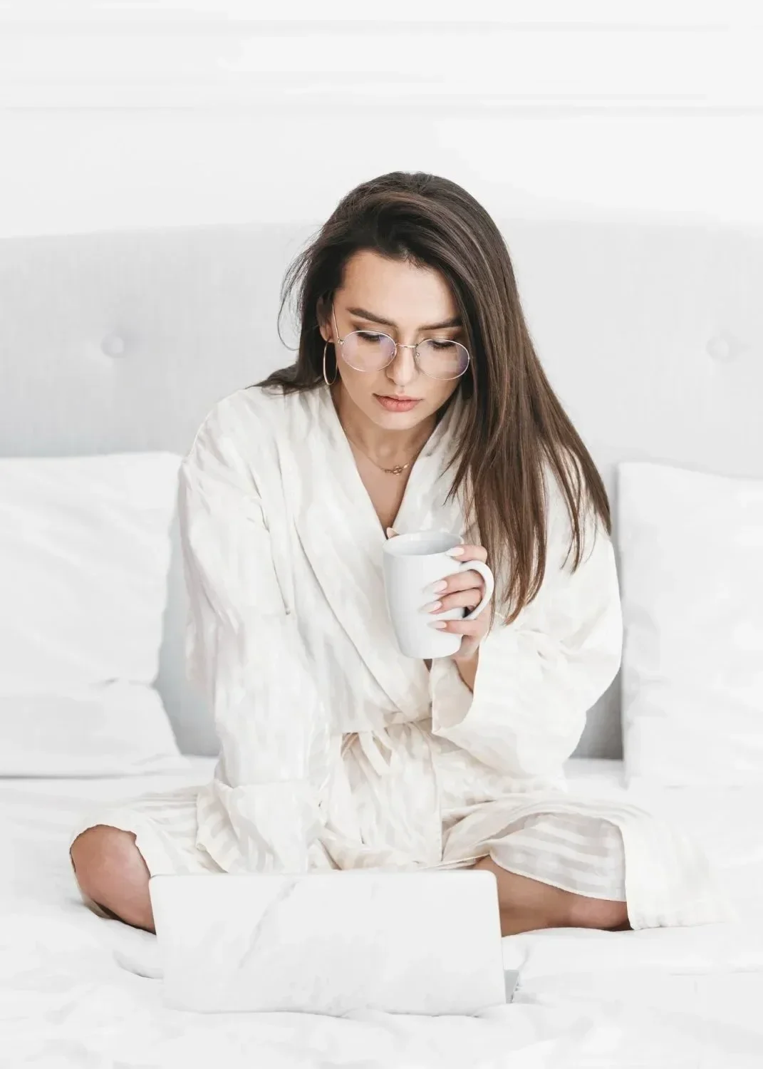 Woman in a white robe sits on a bed with a laptop, holding a mug. She appears focused, creating a cozy and contemplative atmosphere.