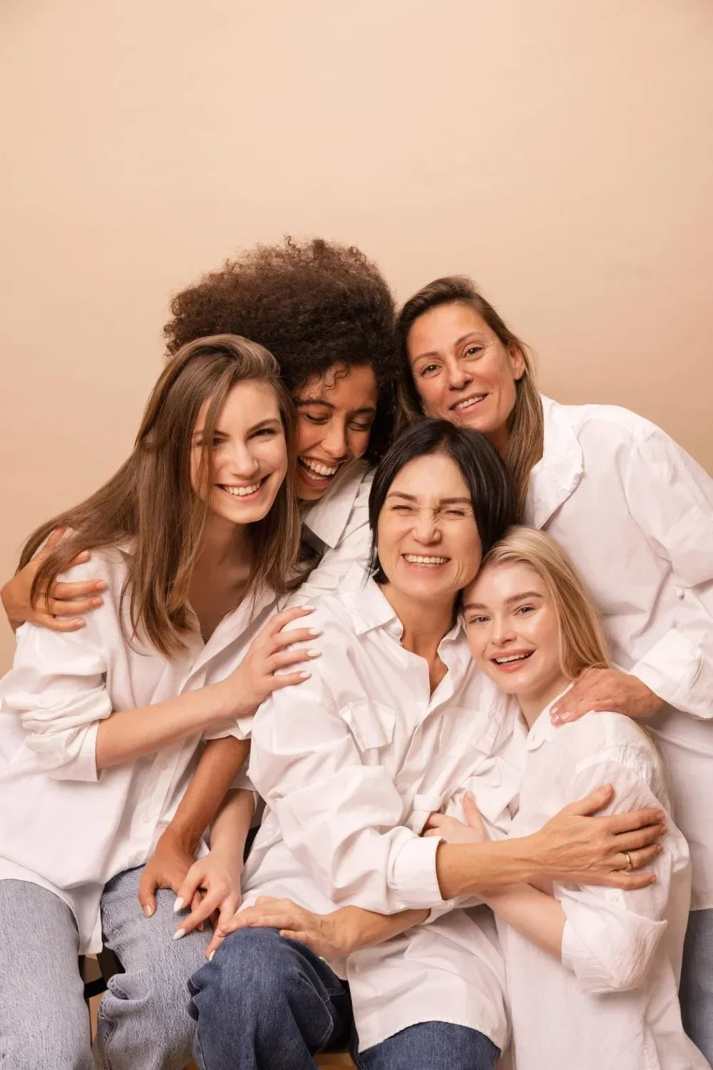 Group of five diverse women wearing white shirts, sitting and standing closely together against a beige background.