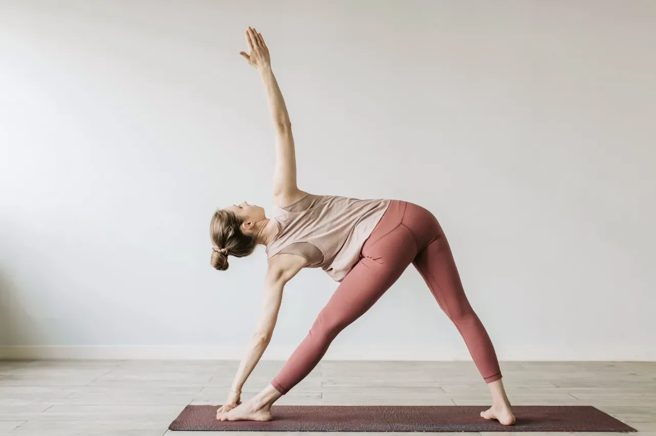 A woman in pink leggings and a beige tank top performs a triangle yoga pose on a brown mat in a bright, minimalist room.