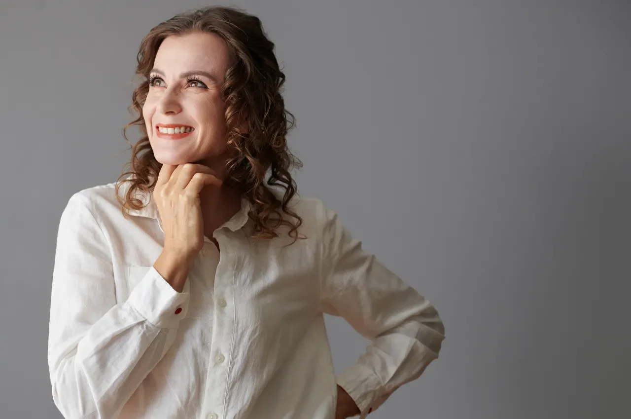 Smiling woman with curly hair wearing a white shirt poses against a gray background. She looks to the side, exuding a thoughtful and cheerful ambiance.