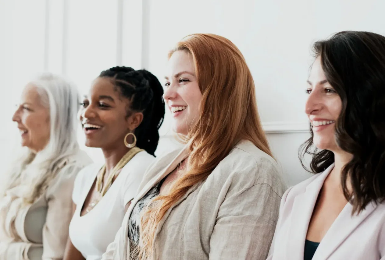 Four women of diverse backgrounds sit in a row, smiling and laughing. They exude happiness and camaraderie in a bright, airy setting.