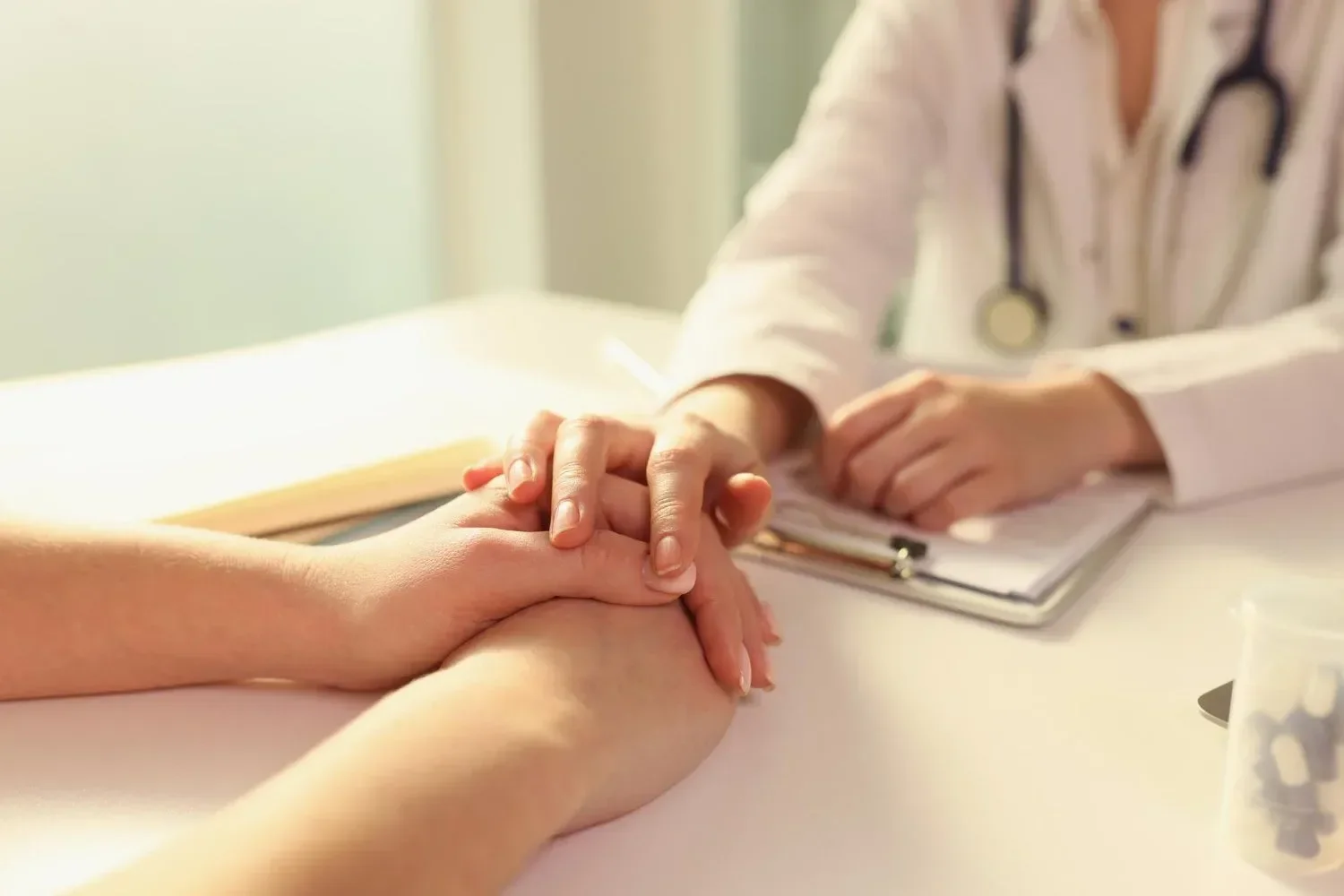 A doctor, wearing a stethoscope, compassionately holds a patient's hand across a desk with medical notes, conveying support and empathy.