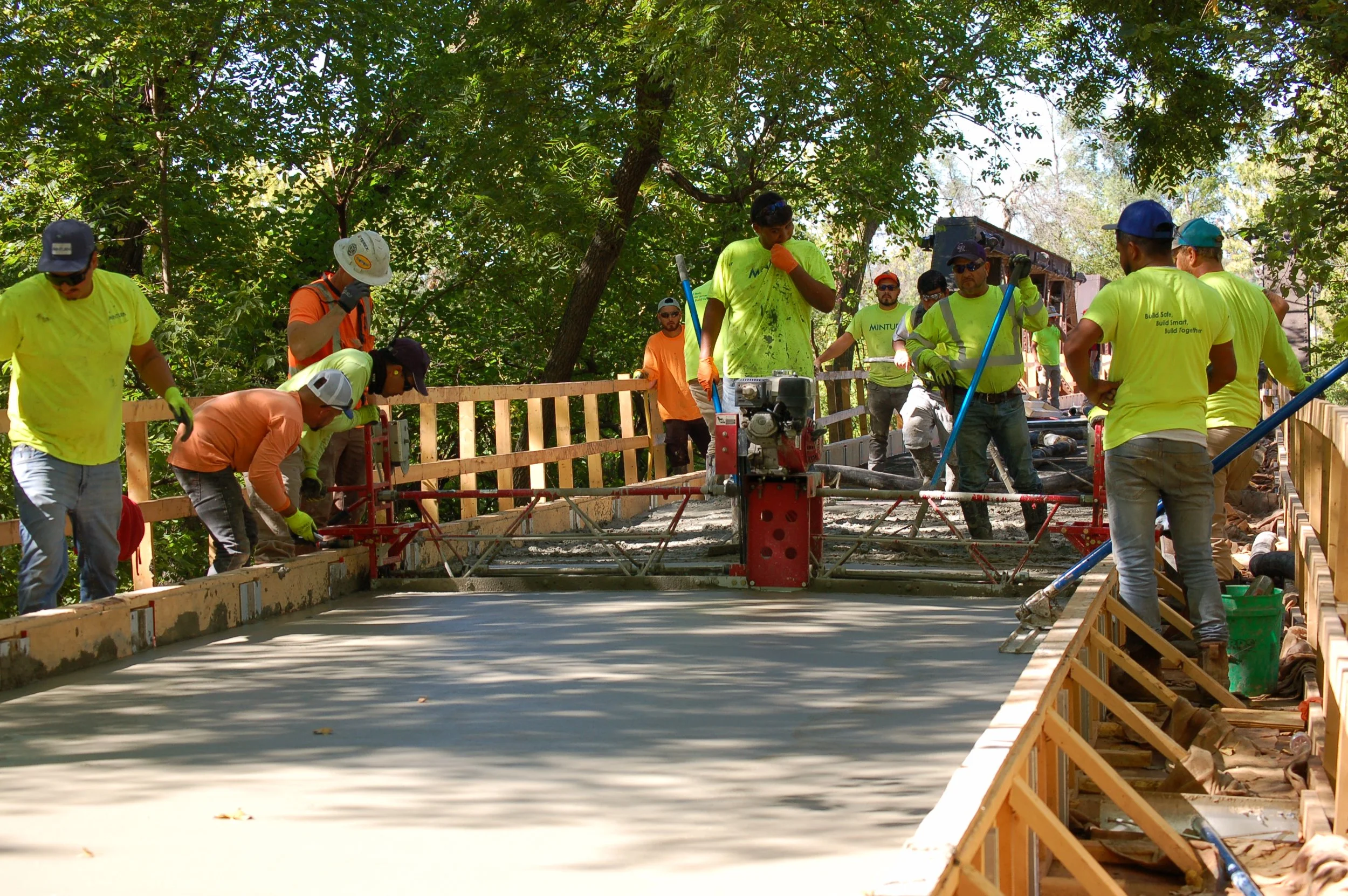 Construction workers smoothing wet concrete on a bridge, surrounded by trees, with wooden safety railing and construction equipment.
