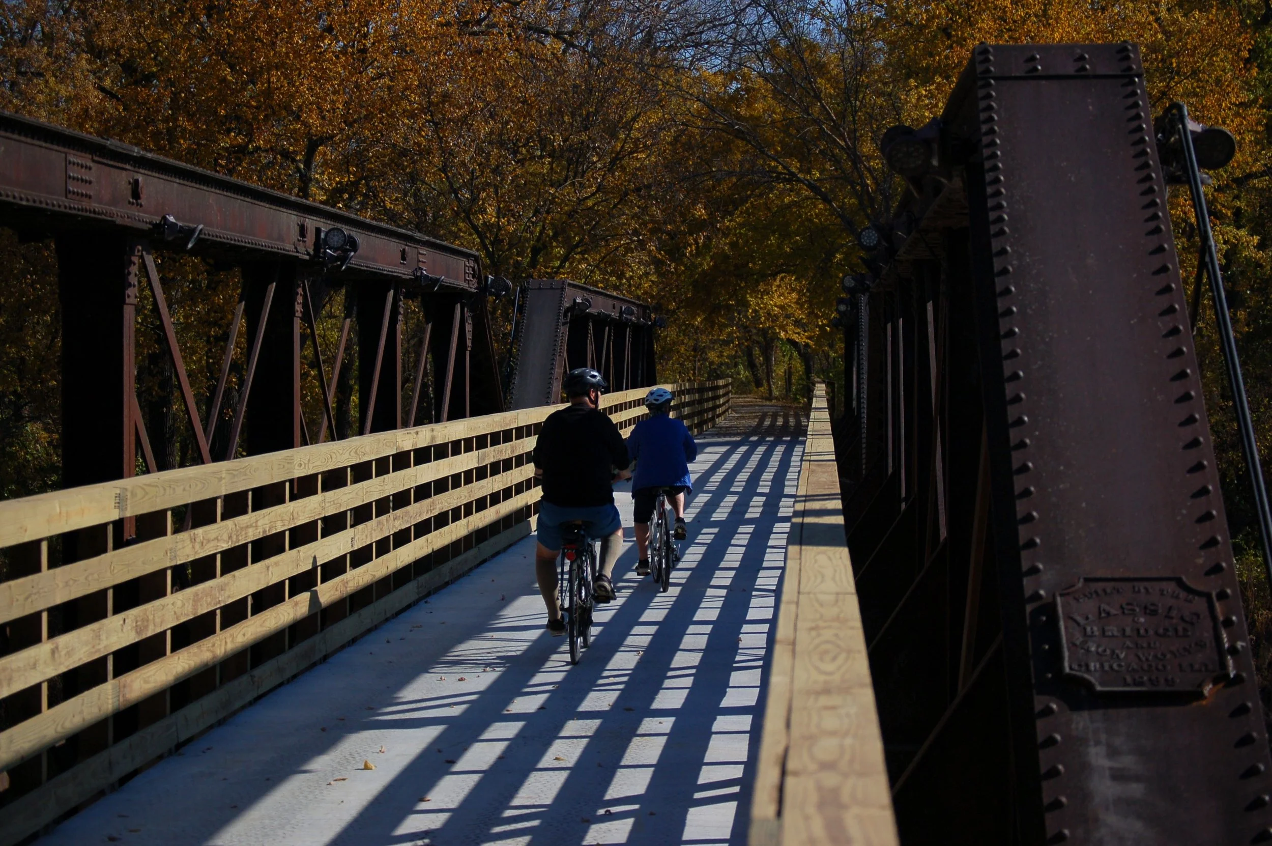 Two people, a man and a child, riding bikes on a narrow bridge during autumn with trees in the background.