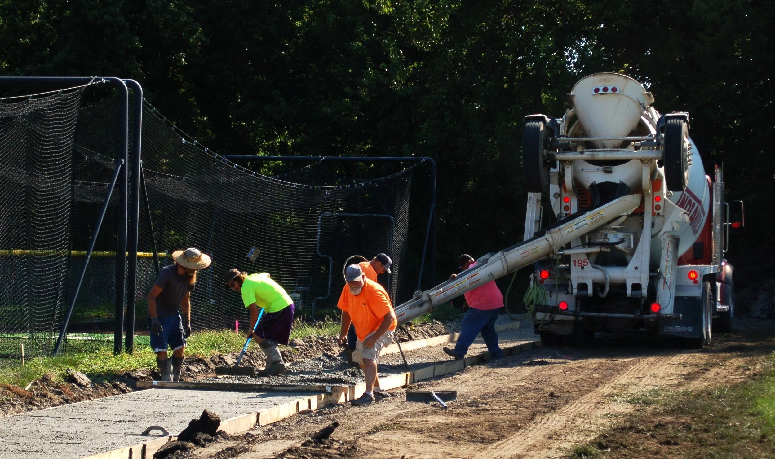 Workers laying concrete on a sidewalk with a cement mixer truck nearby, some workers wearing hats and bright shirts, working along a fence with trees in the background.
