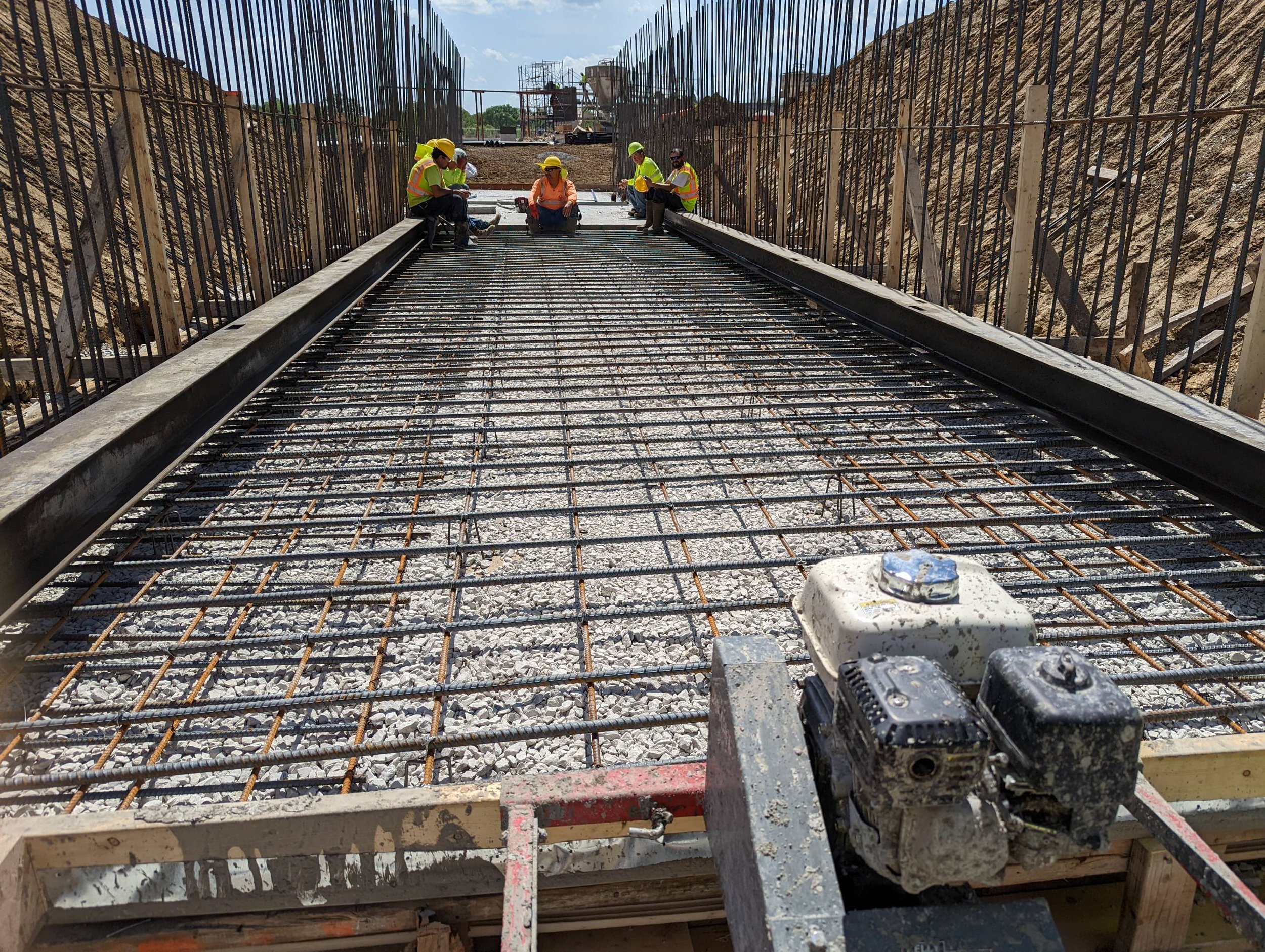 Construction workers in yellow safety vests and helmets working on concrete reinforcement formwork for a football stadium tunnel under a blue sky.