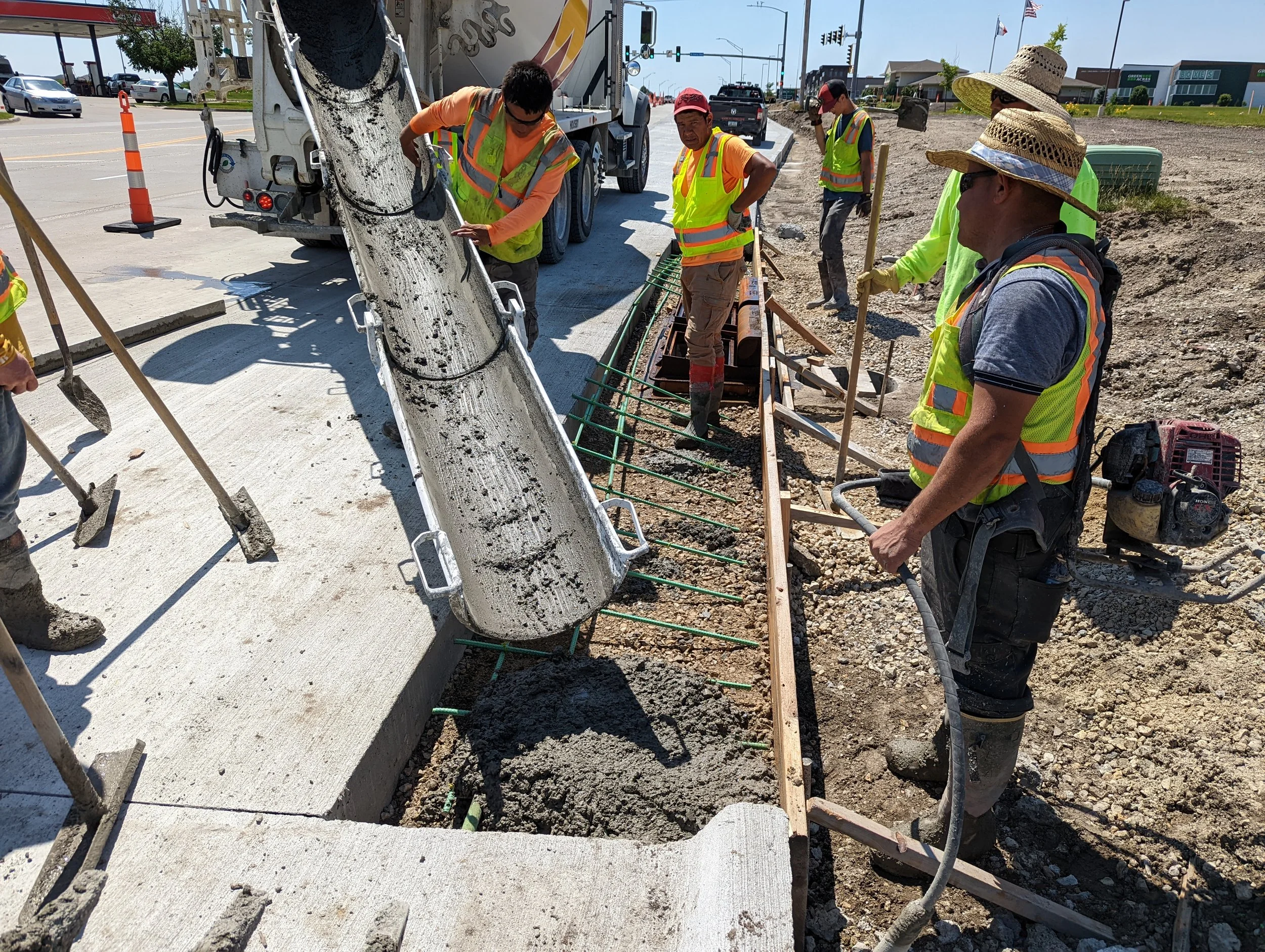 Construction workers pouring concrete into a form reinforced with steel rebar at a sidewalk or curb site on a sunny day.