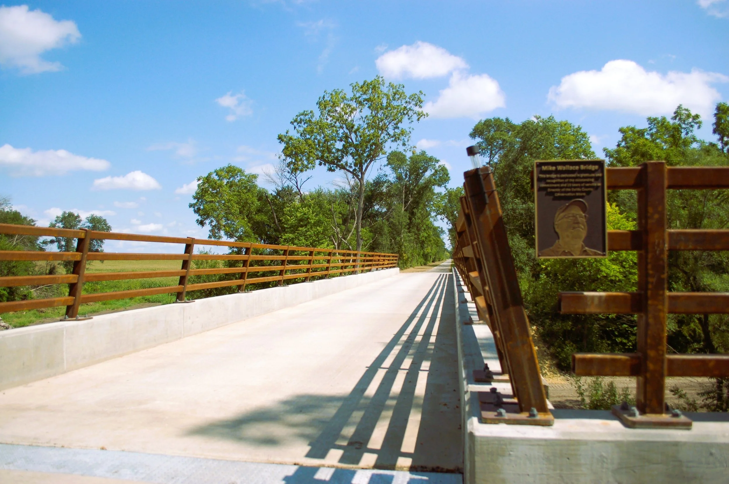A bridge with steel railing on both sides, surrounded by green trees and a bright blue sky with scattered clouds.